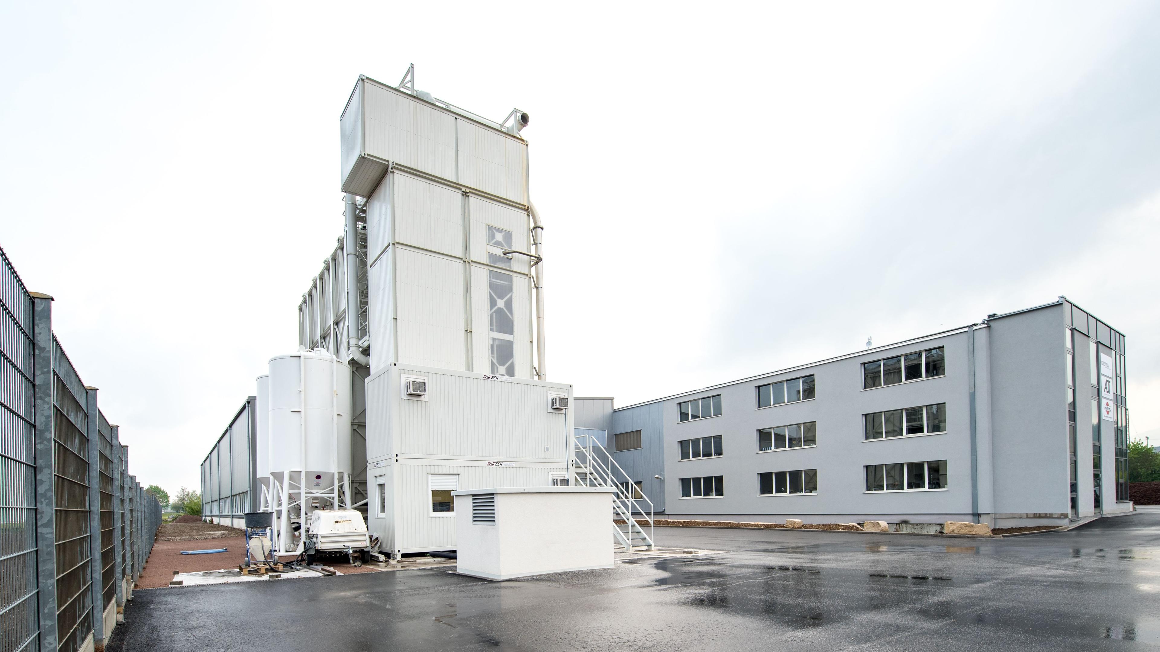 Industrieanlage mit großen weißen Silos und angrenzendem grauen Bürogebäude, umgeben von einem Zaun an einem nassen, bewölkten Tag.