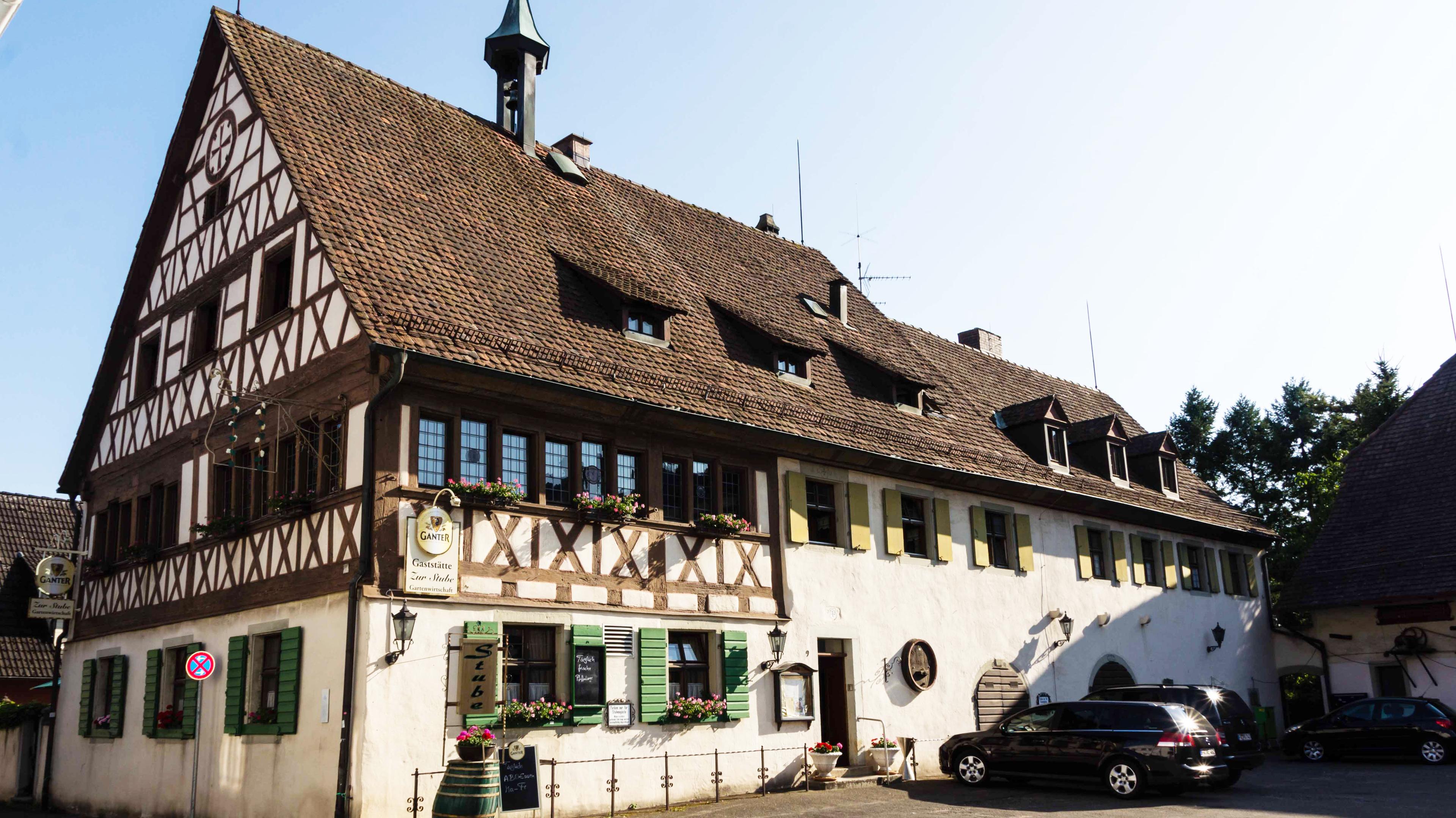 Traditionelles Fachwerkhaus mit steilem Dach, Blumenkästen und geparkten Autos davor, unter strahlend blauem Himmel.