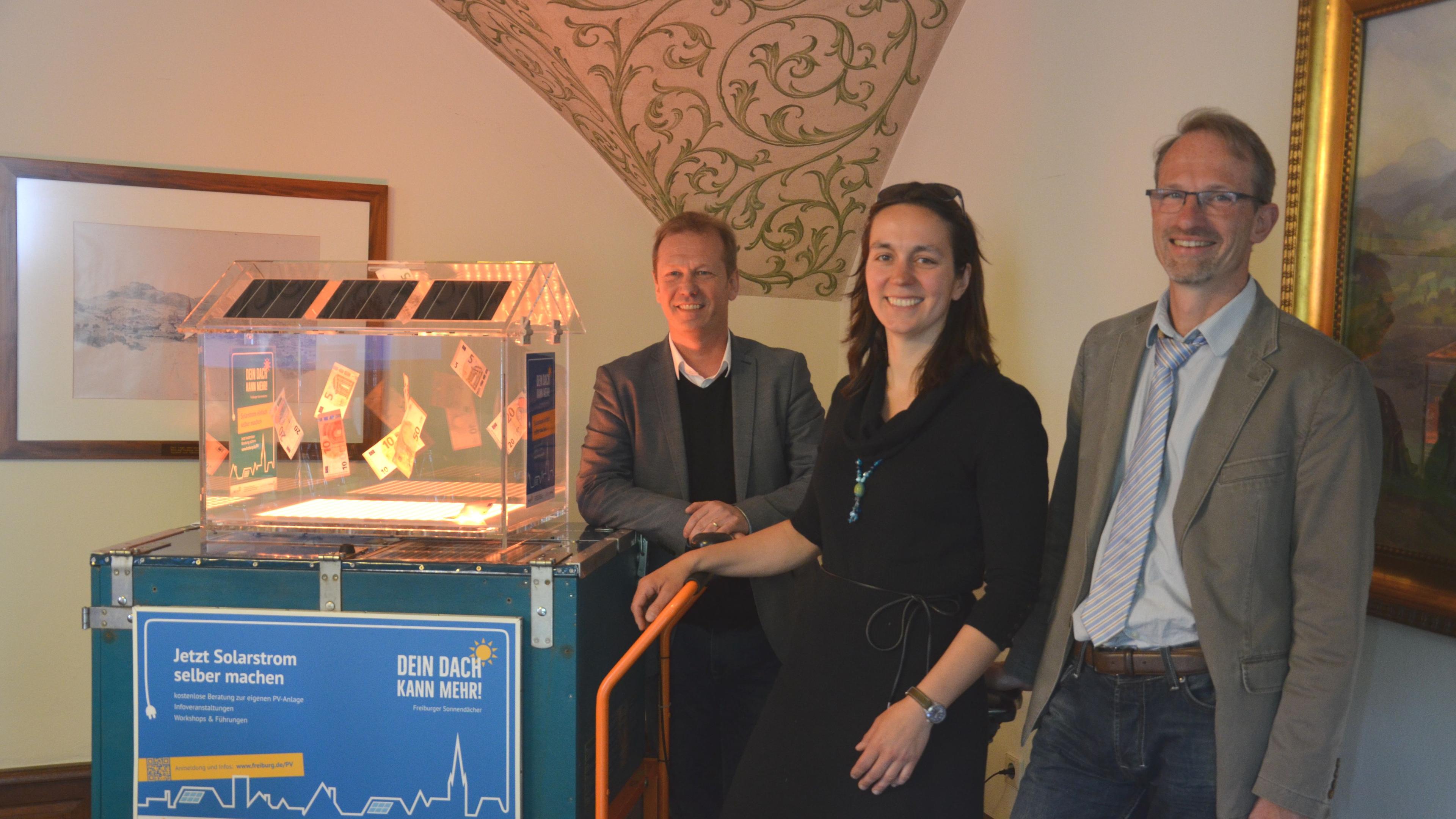 Drei Personen stehen neben einem Solarenergie-Modell auf einem Wagen, neben dem ein Schild für Solarenergie steht. Der Raum ist mit Kunstwerken und kunstvollen Dekorationen ausgestattet.