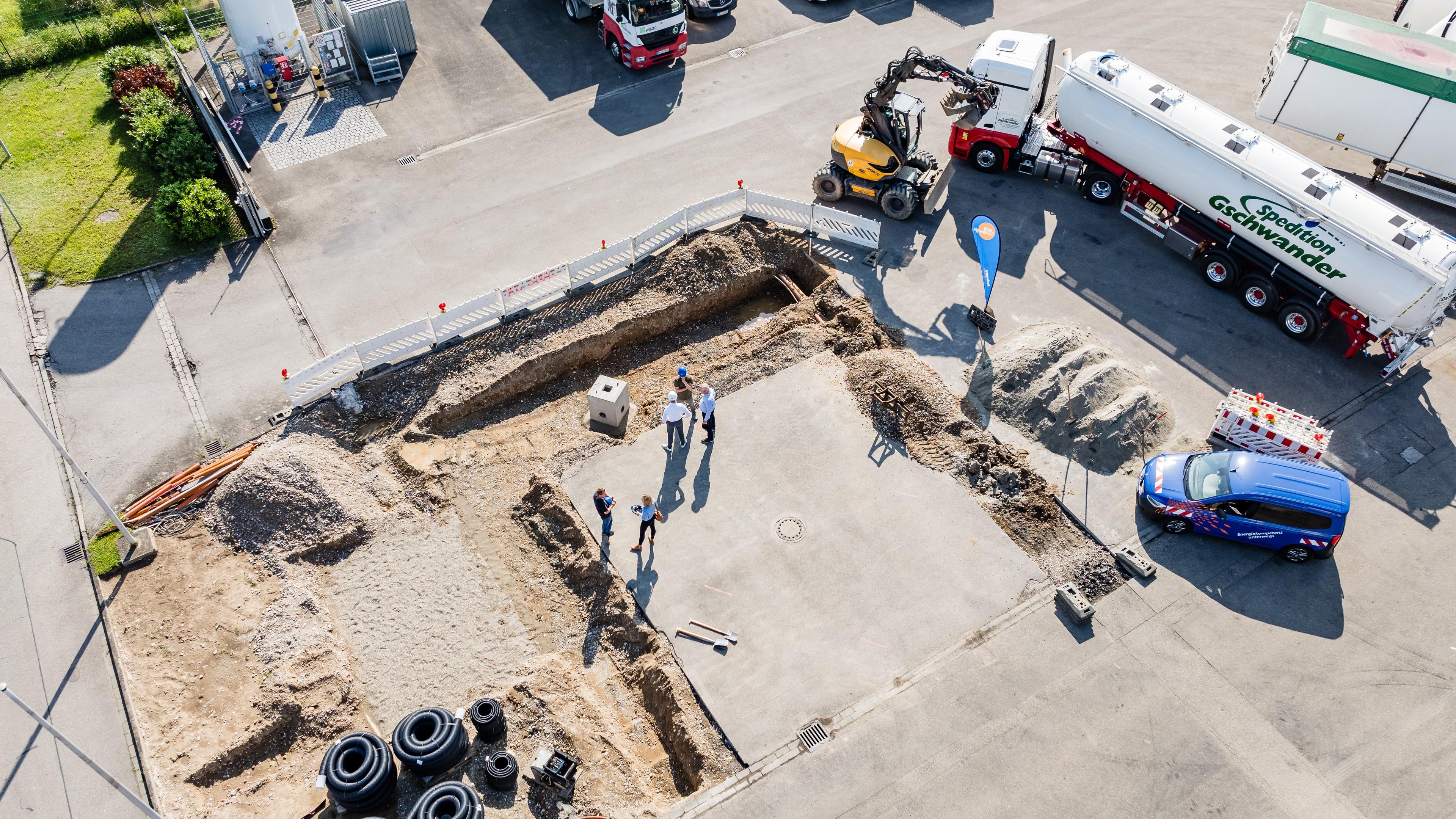 Luftansicht einer Bausteller mit Fahrzeugen, die darum herum parken und Personen an der Baustelle, die sich unterhalten.