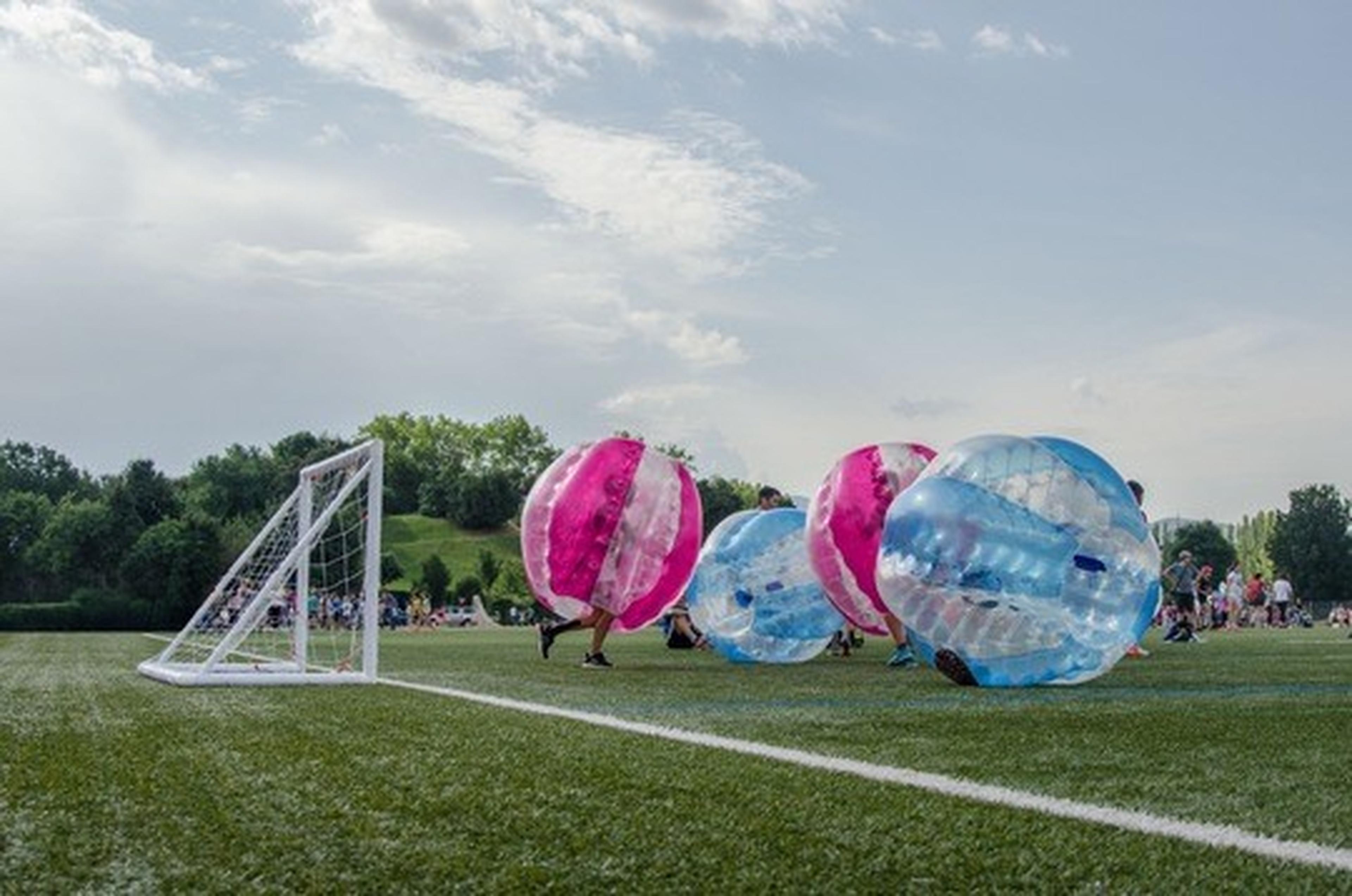 People in colorful inflatable bubbles playing soccer on a grassy field, near a goalpost, with trees and people in the background.
