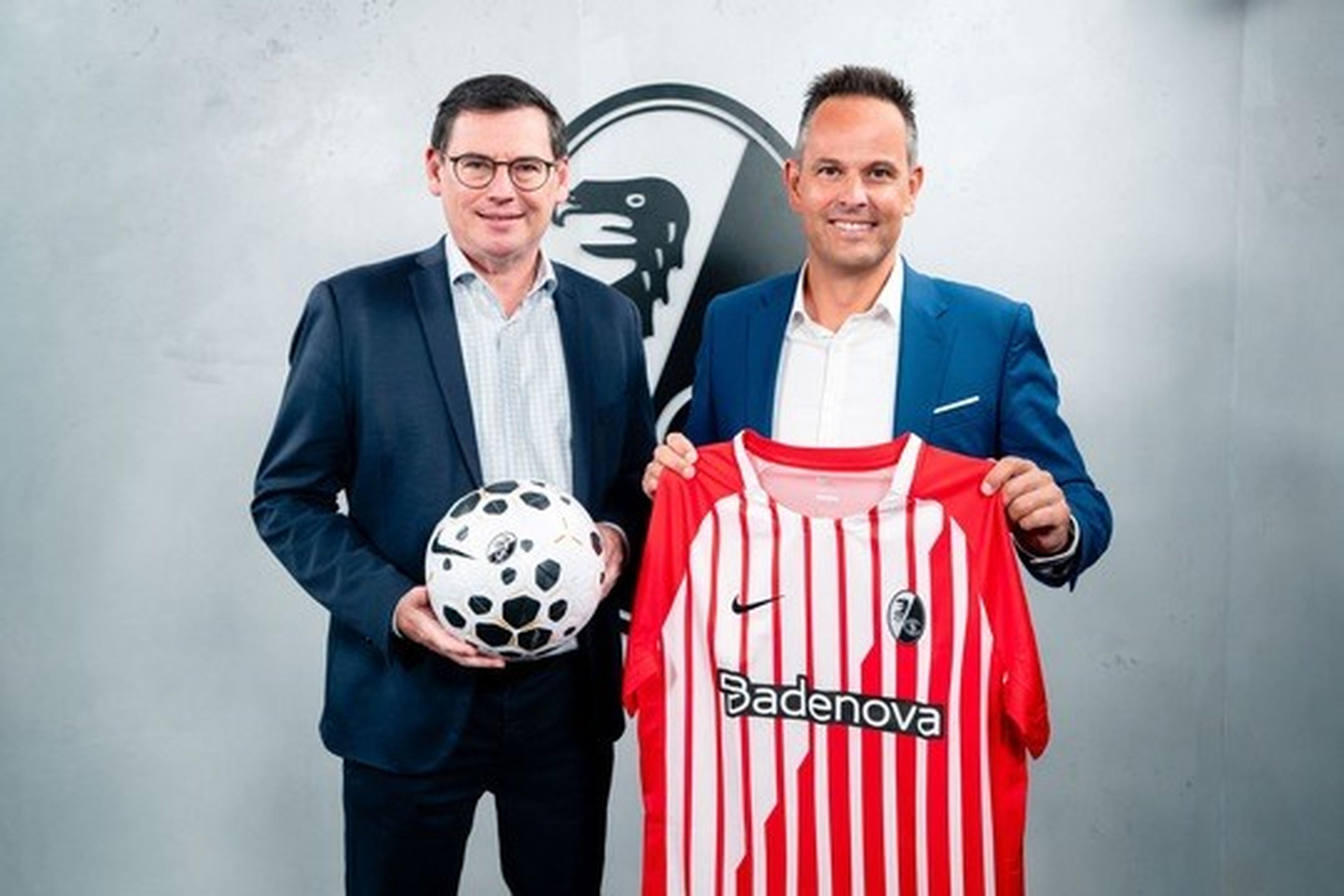 Two men in suits; one holding a soccer ball, the other displaying a red and white striped soccer jersey with the Badenova logo.