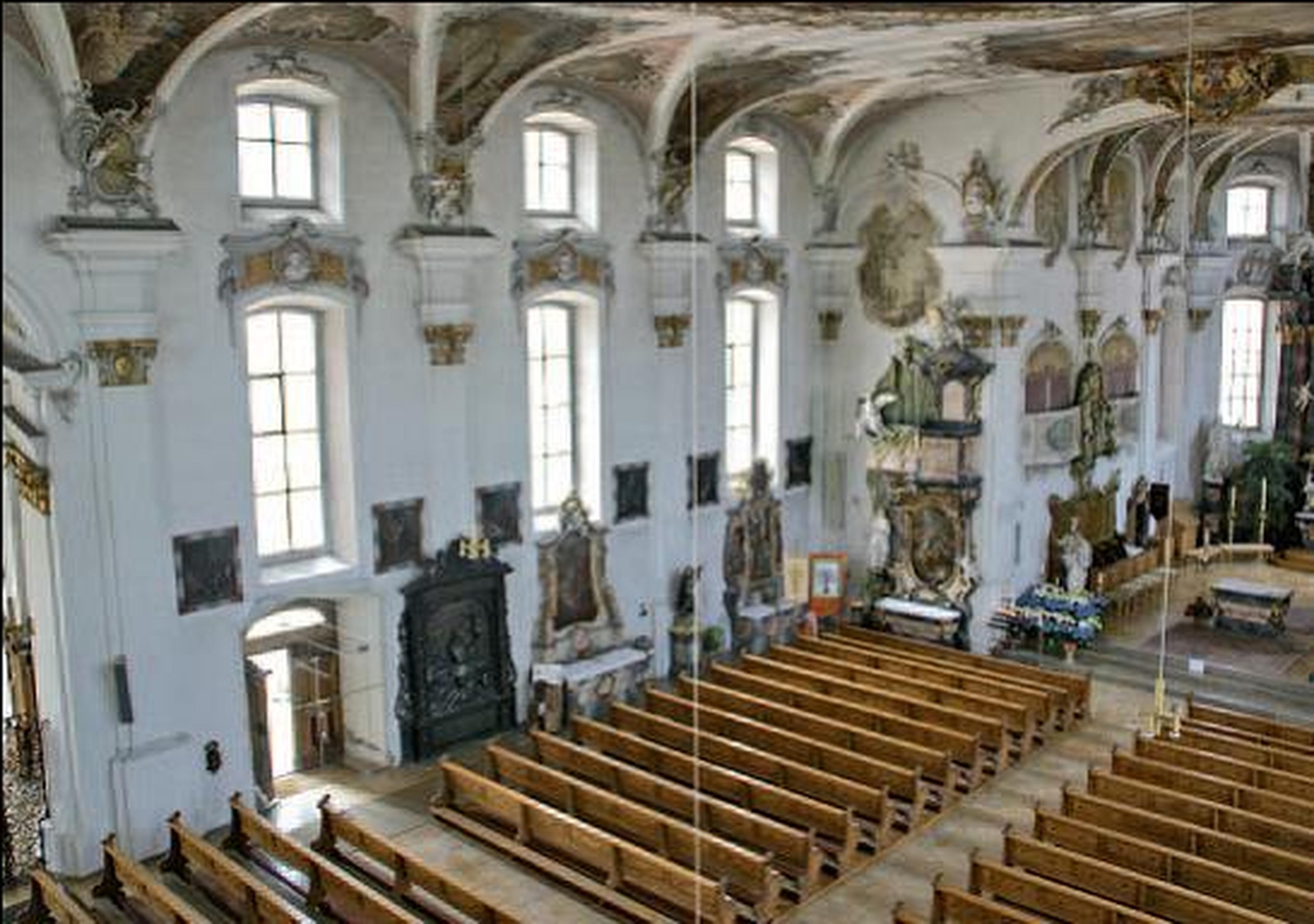 Das Innere einer Barockkirche mit hohen Bogenfenstern, kunstvollen Fresken an der Decke, Holzbänken und aufwendigen Altarverzierungen.