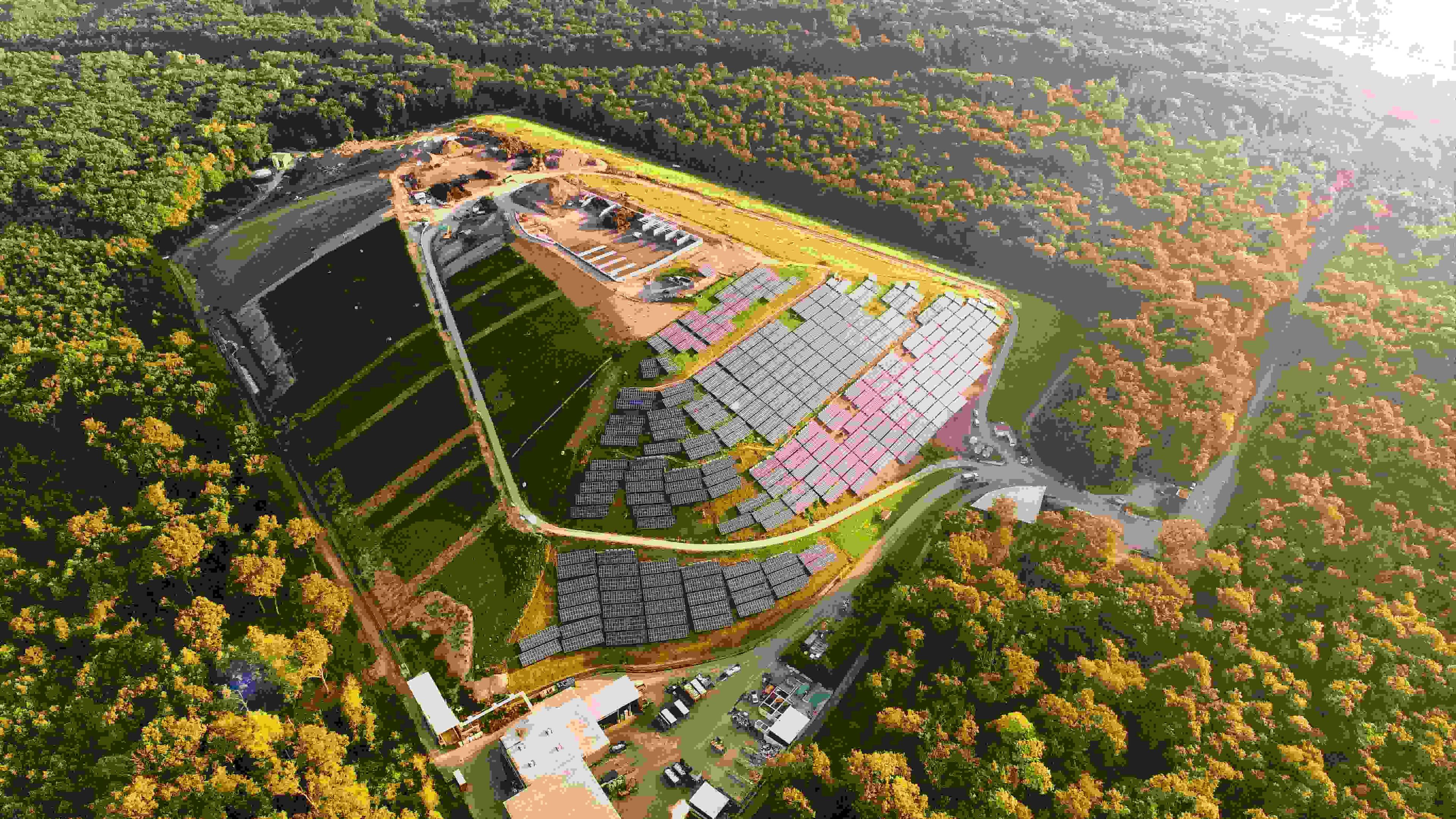 Luftaufnahme einer Solarfarm, umgeben von dichtem, farbenfrohem Herbstwald, mit in Reihen angeordneten Solarmodulen und sichtbarer Konstruktion im oberen Bereich.