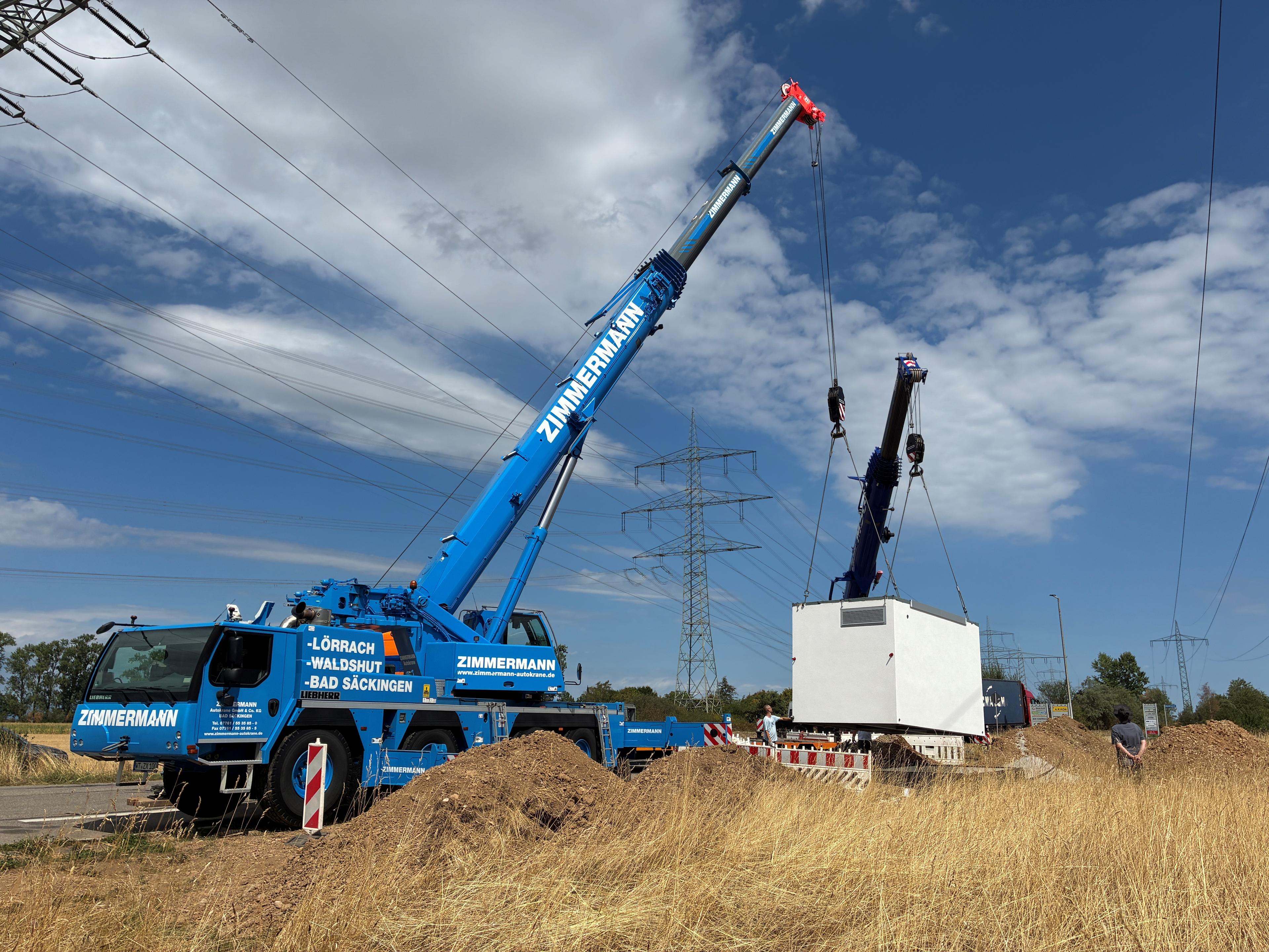 Two blue cranes lift a large white container on a construction site with clear skies and power lines in the background.