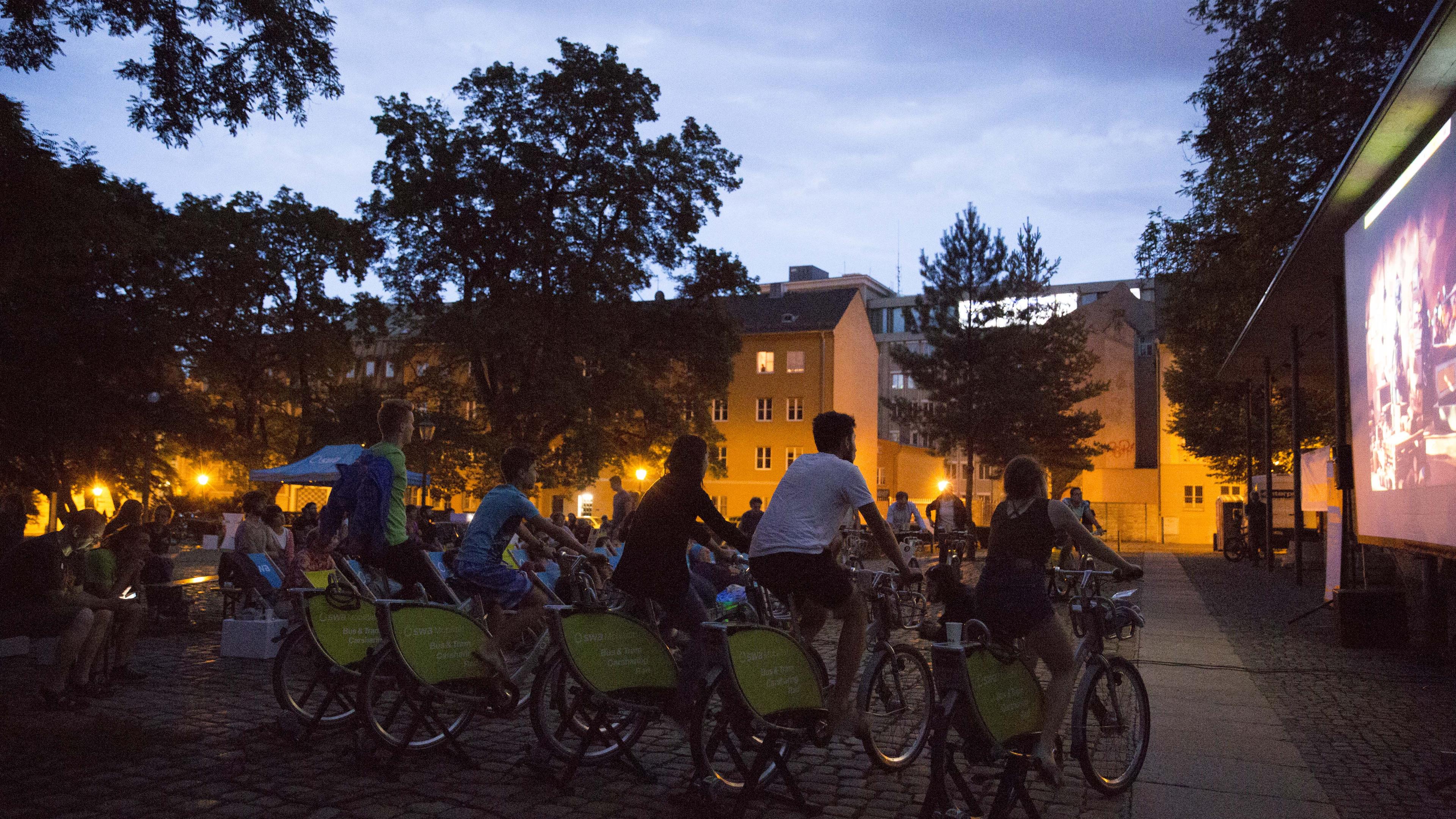 Menschen auf stationären Fahrrädern im Freien schauen sich in der Abenddämmerung einen Film an, der auf eine Leinwand projiziert wird, umgeben von Bäumen und beleuchteten Gebäuden.