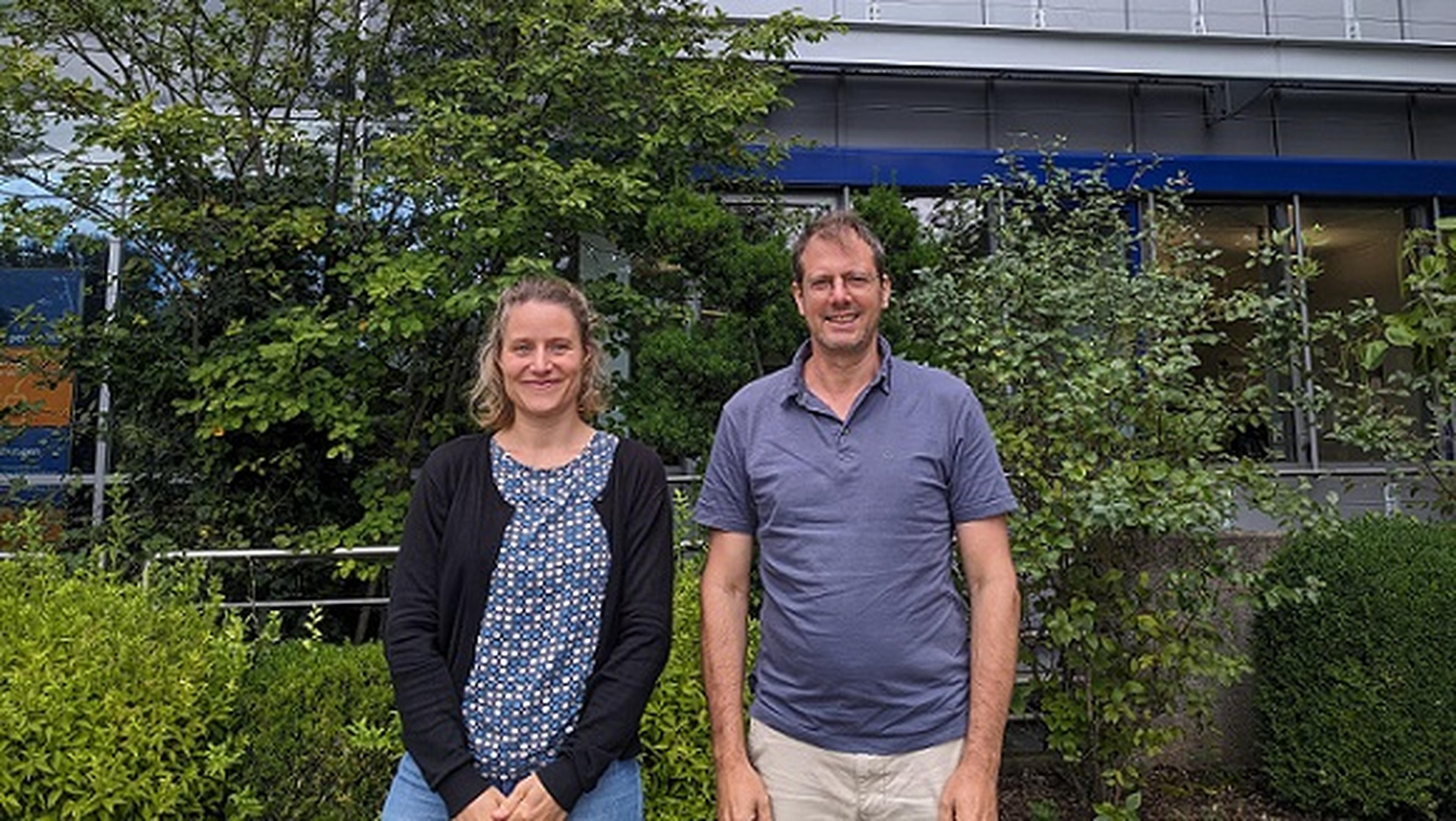 Zwei Personen stehen vor einem Gebäude mit Grünpflanzen und lächeln in die Kamera.