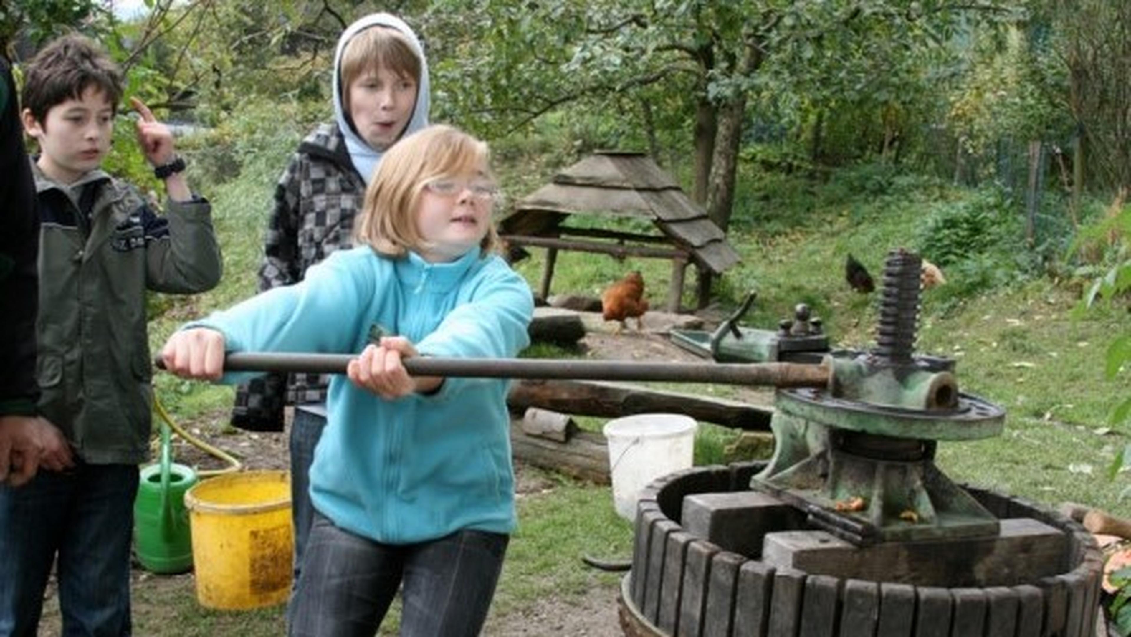 Three children outdoors, with one using a vintage cider press. Chickens and buckets are in the background, surrounded by greenery.