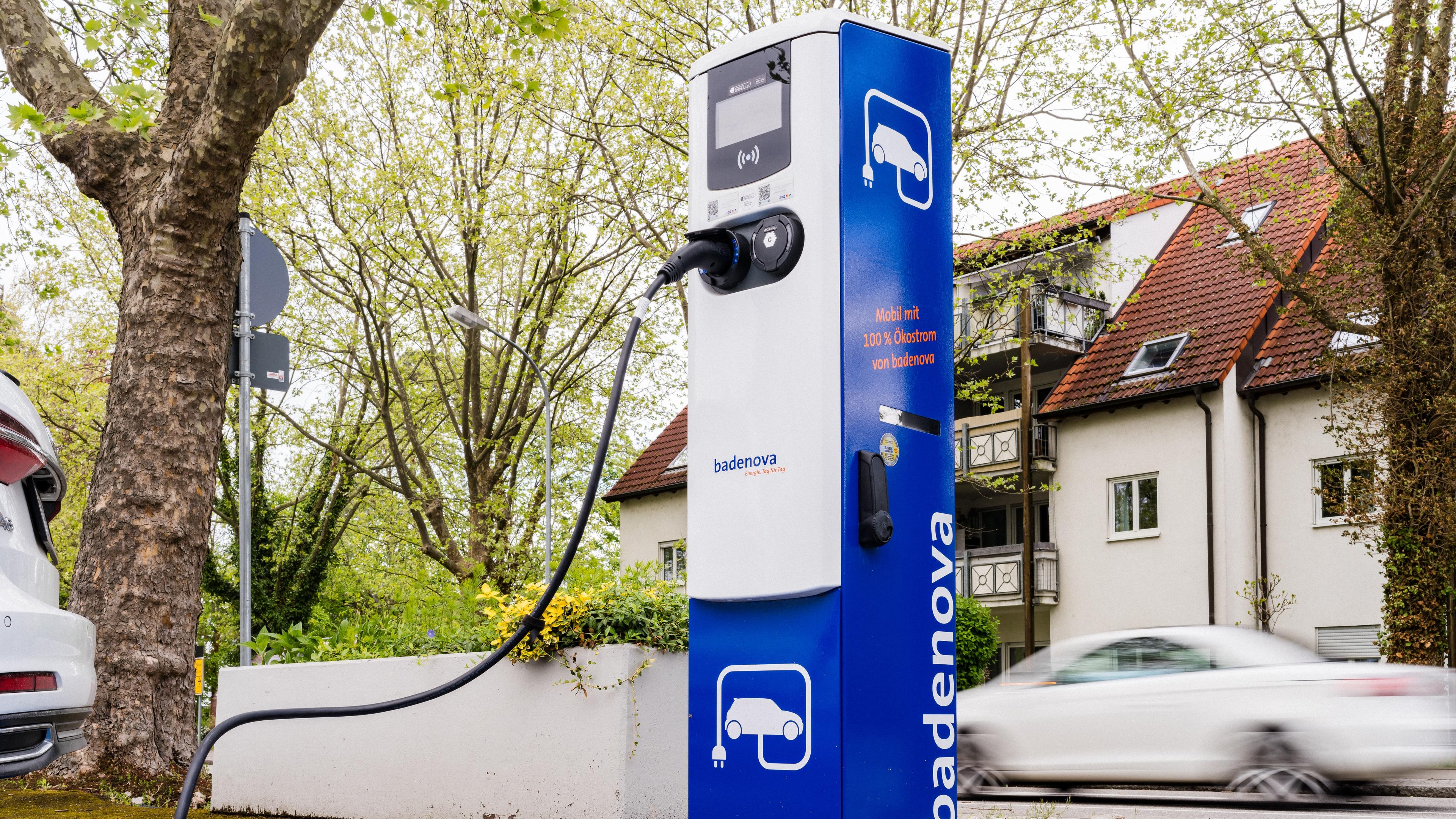 Ladesäule mit eingestecktem Ladekabel vor einer befahrenen Straße in einer Ortschaft.
