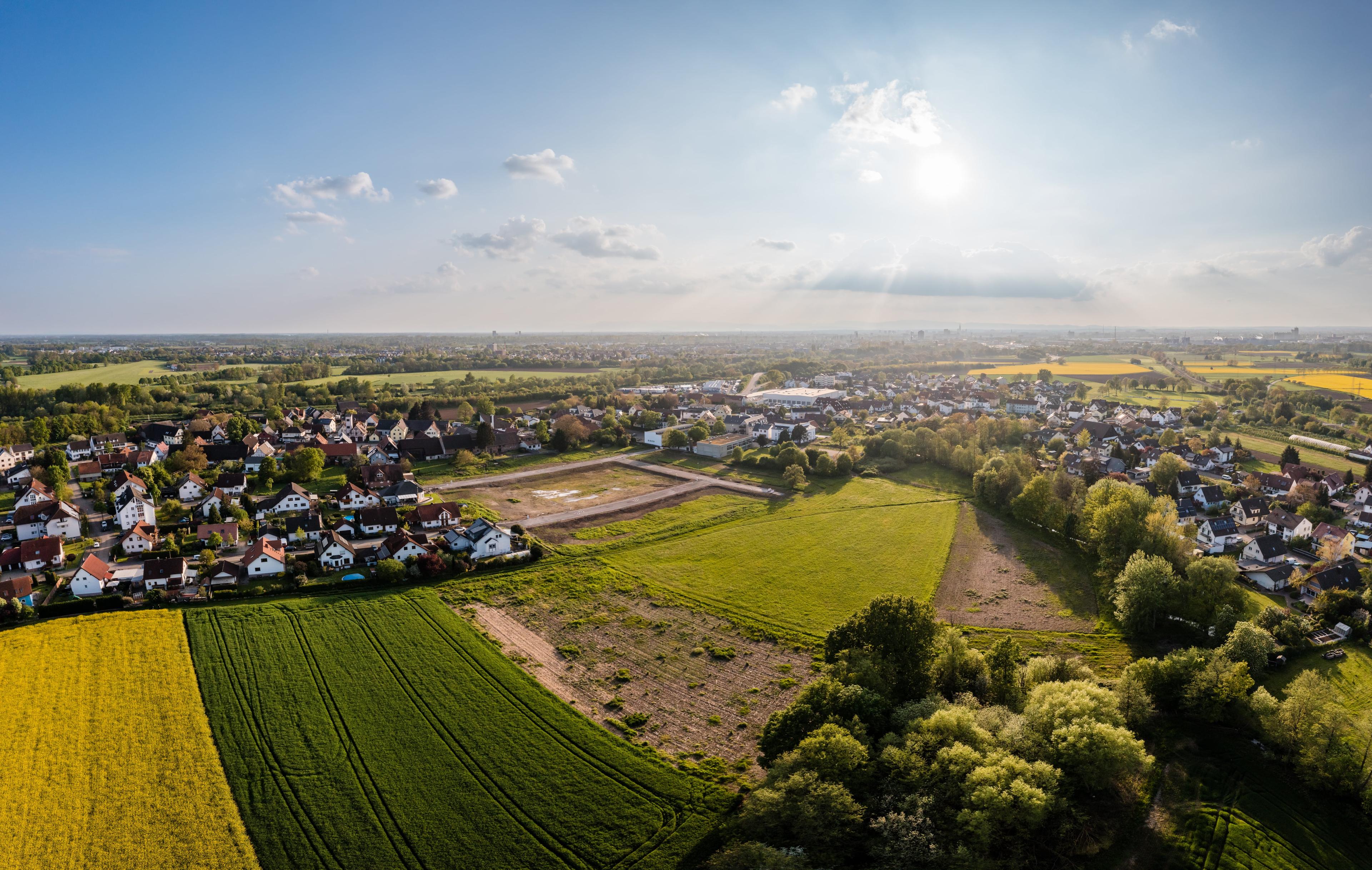 Luftaufnahme einer ländlichen Landschaft mit Feldern, Bäumen und einer Häusergruppe unter blauem Himmel mit vereinzelten Wolken und Sonnenschein.