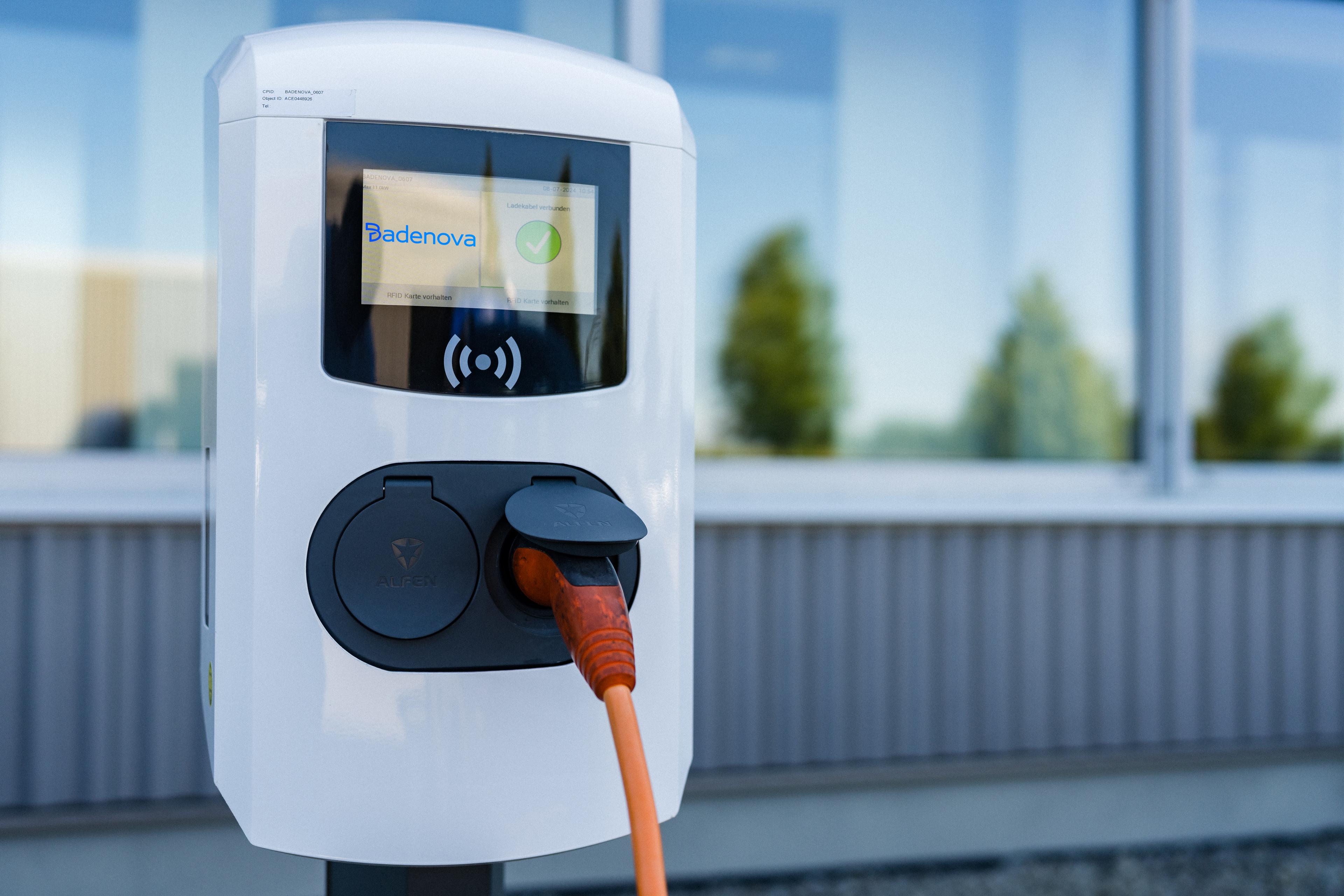 Aufgeständerte Wallbox mit eingestecktem orangenen Ladekabel im Vordergrund eines Gewerbegebäudes.