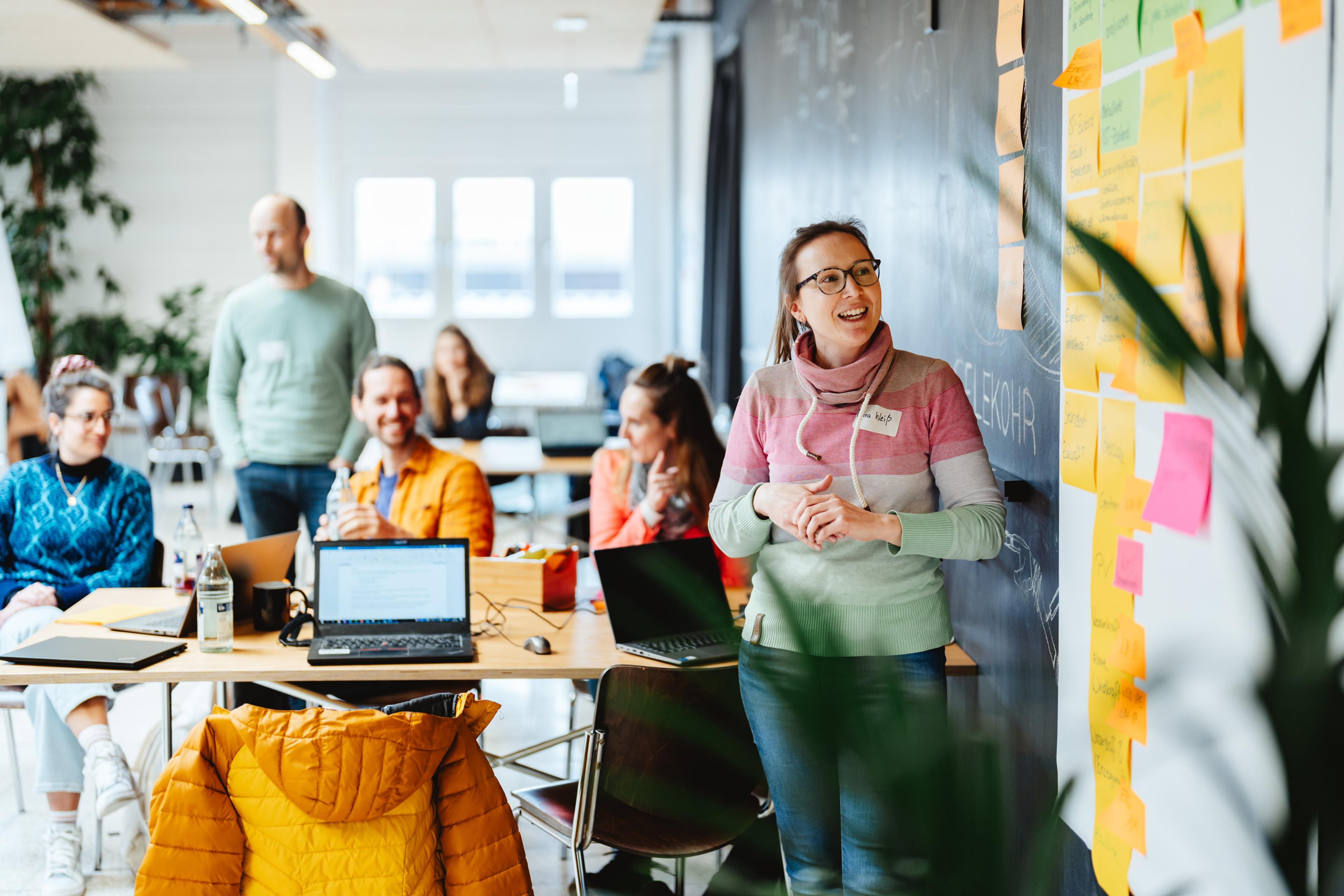 Eine Frau hält einen Vortrag in einem Workshop und steht dabei vor einer Wand voller Haftnotizen. Die Teilnehmer sitzen an Tischen mit Laptops, sind aufmerksam und hören zu.