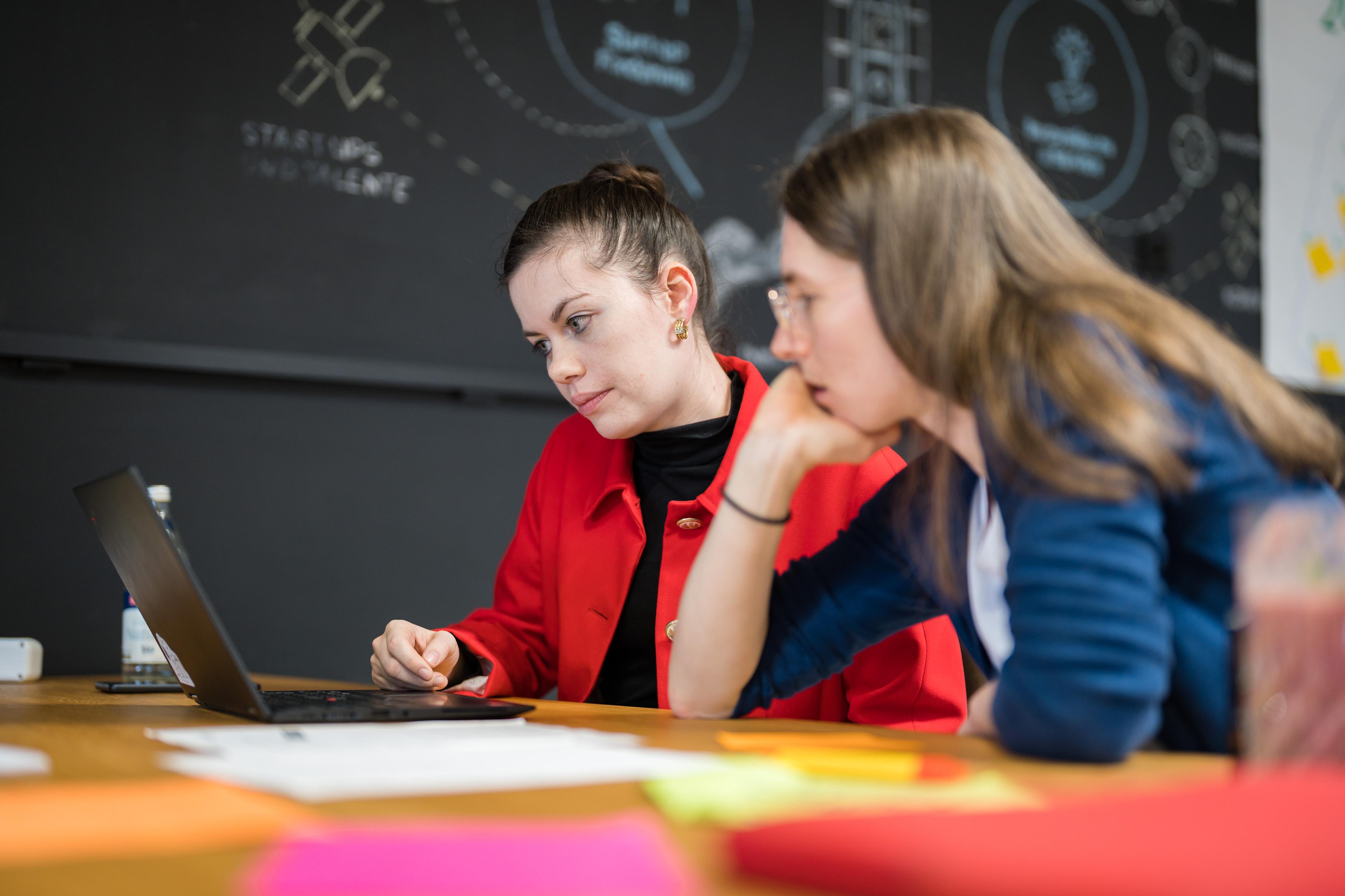 Zwei Frauen, die auf einen Laptop-Bildschirm an einem Schreibtisch mit bunten Papieren konzentriert sind, vor einer Tafel mit Diagrammen im Hintergrund.