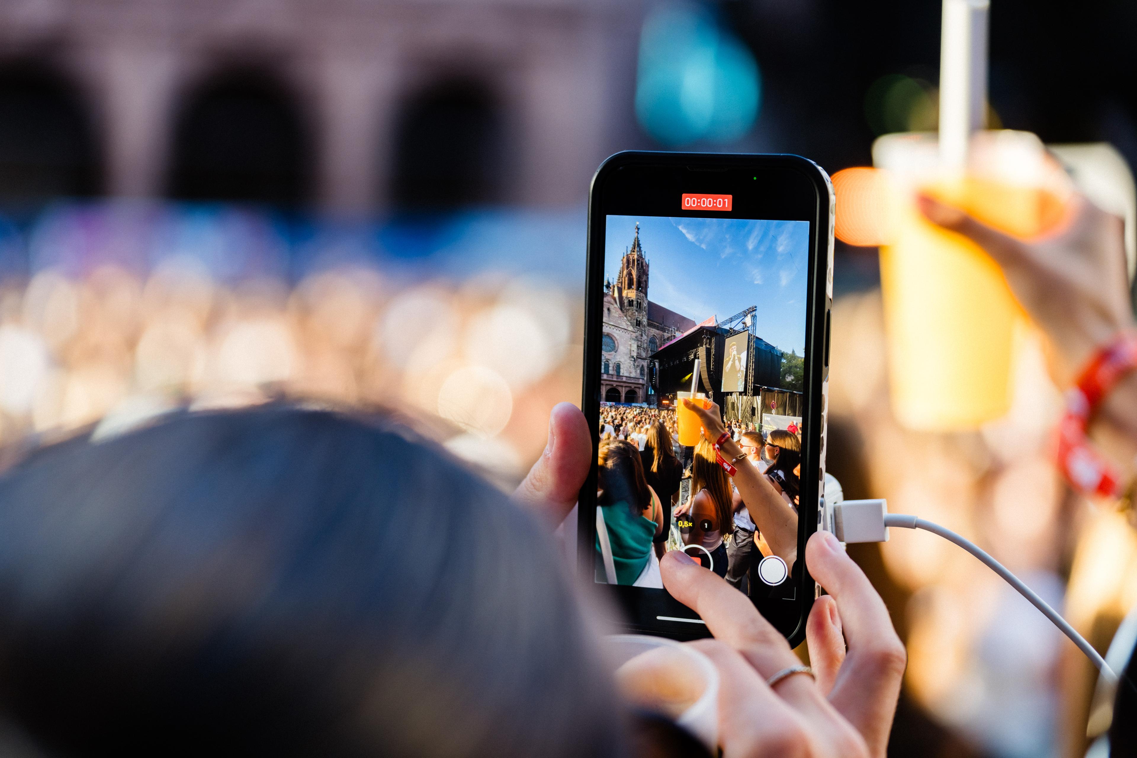 Person, die mit einem Smartphone ein lebhaftes Open-Air-Konzert auf dem Münsterplatz in Freiburg  aufzeichnet und dabei die Bühne und das Publikum einfängt. Helle, sonnige Atmosphäre mit unscharfem Hintergrund.