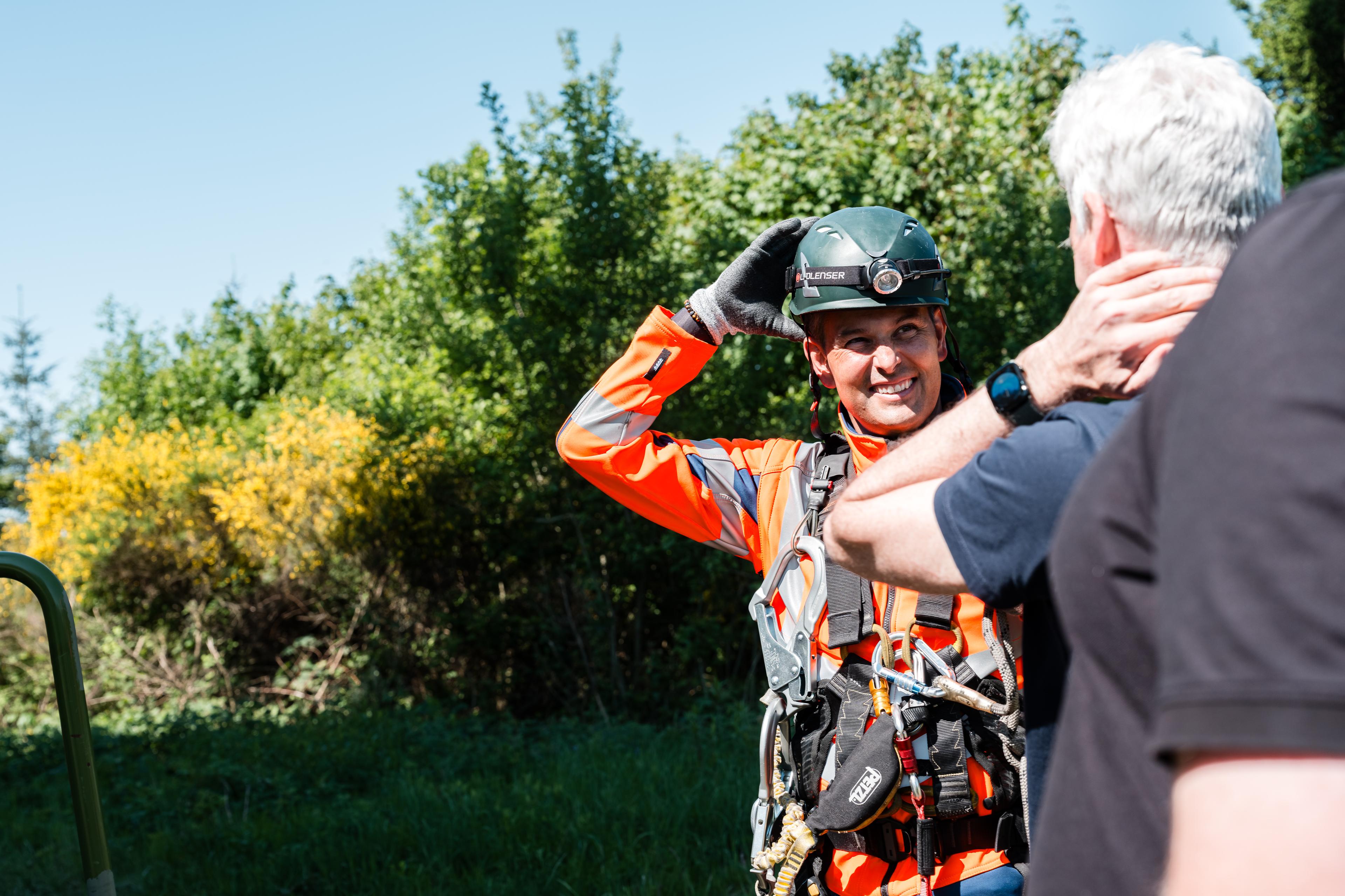 Ein lächelnder Arbeiter in orangefarbener Warnkleidung und mit Sicherheitsgurt, der seinen Helm zurechtzieht, während er sich im Freien zwischen grünen Bäumen mit einem Kollegen unterhält