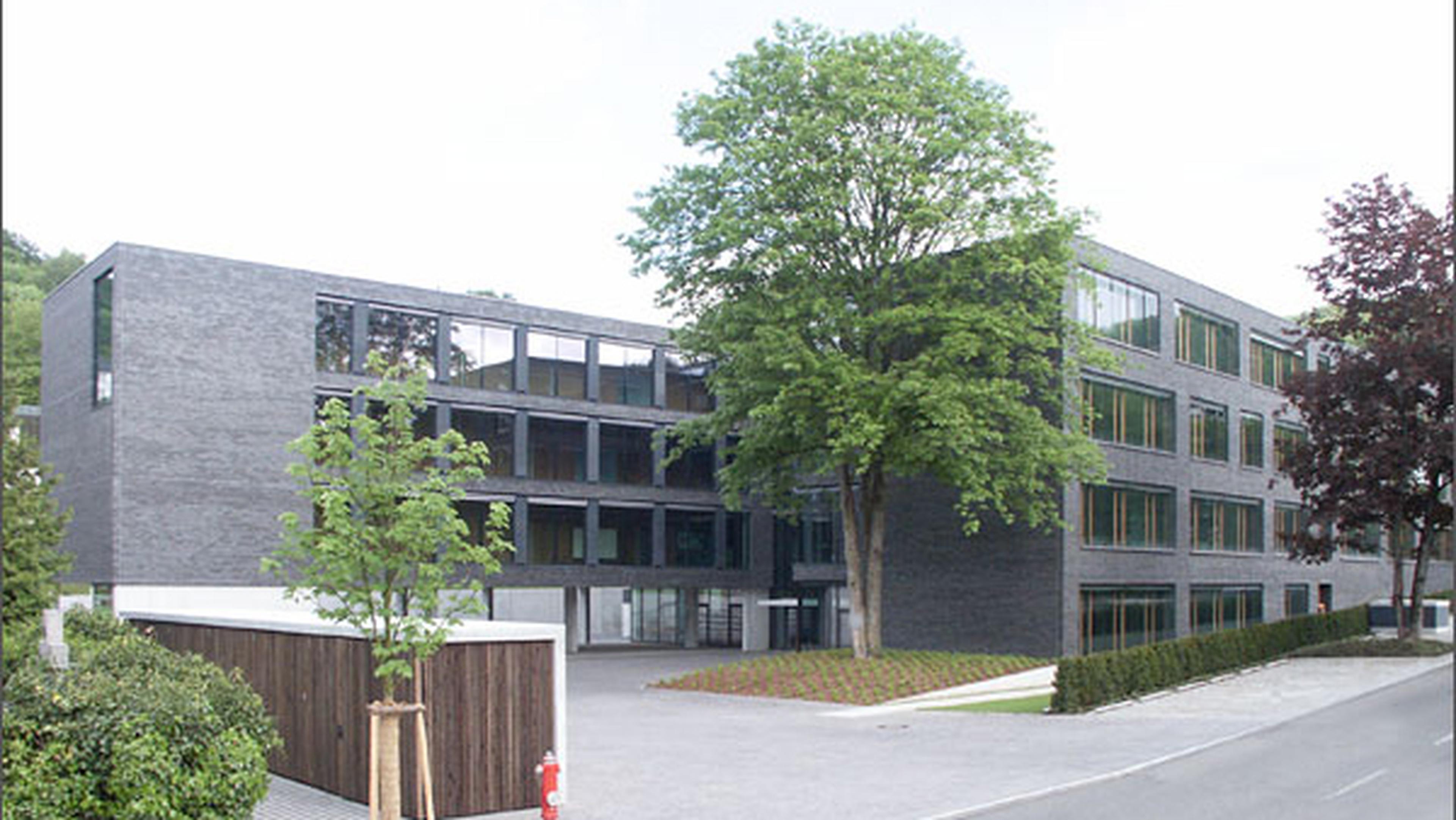 Modernes Bürogebäude mit einer dunklen Backsteinfassade, großen Fenstern und umgeben von Bäumen und viel Grün.