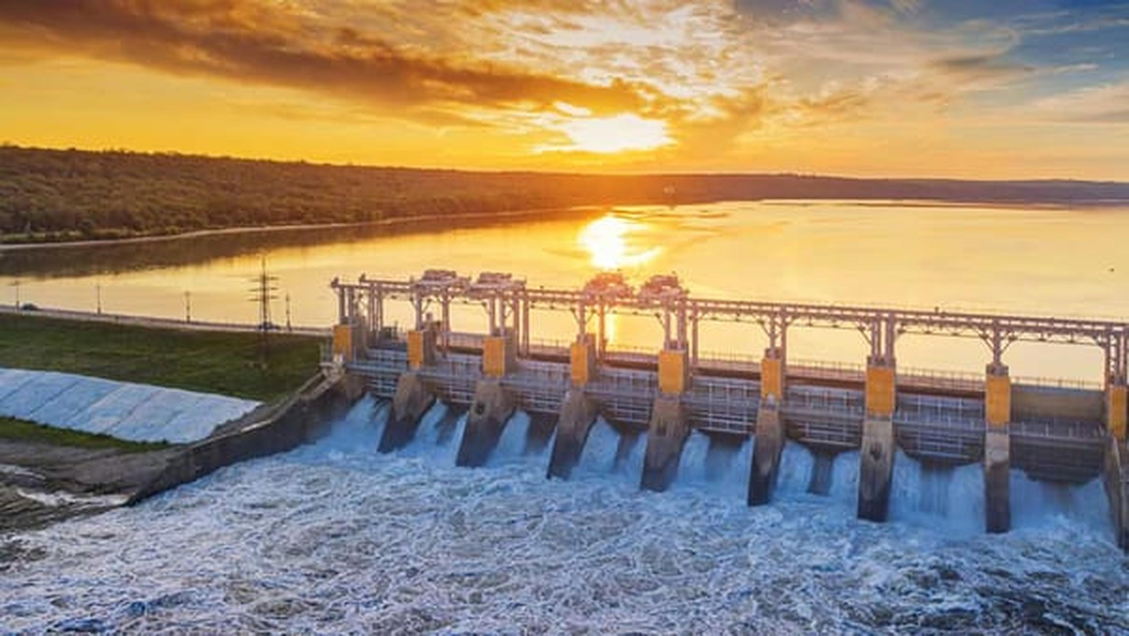 Wasserkraftwerk mit Wasser, das bei Sonnenuntergang durch Schleusen fließt, umgeben von einem Wald und einem riesigen See, der den farbenfrohen Himmel widerspiegelt.