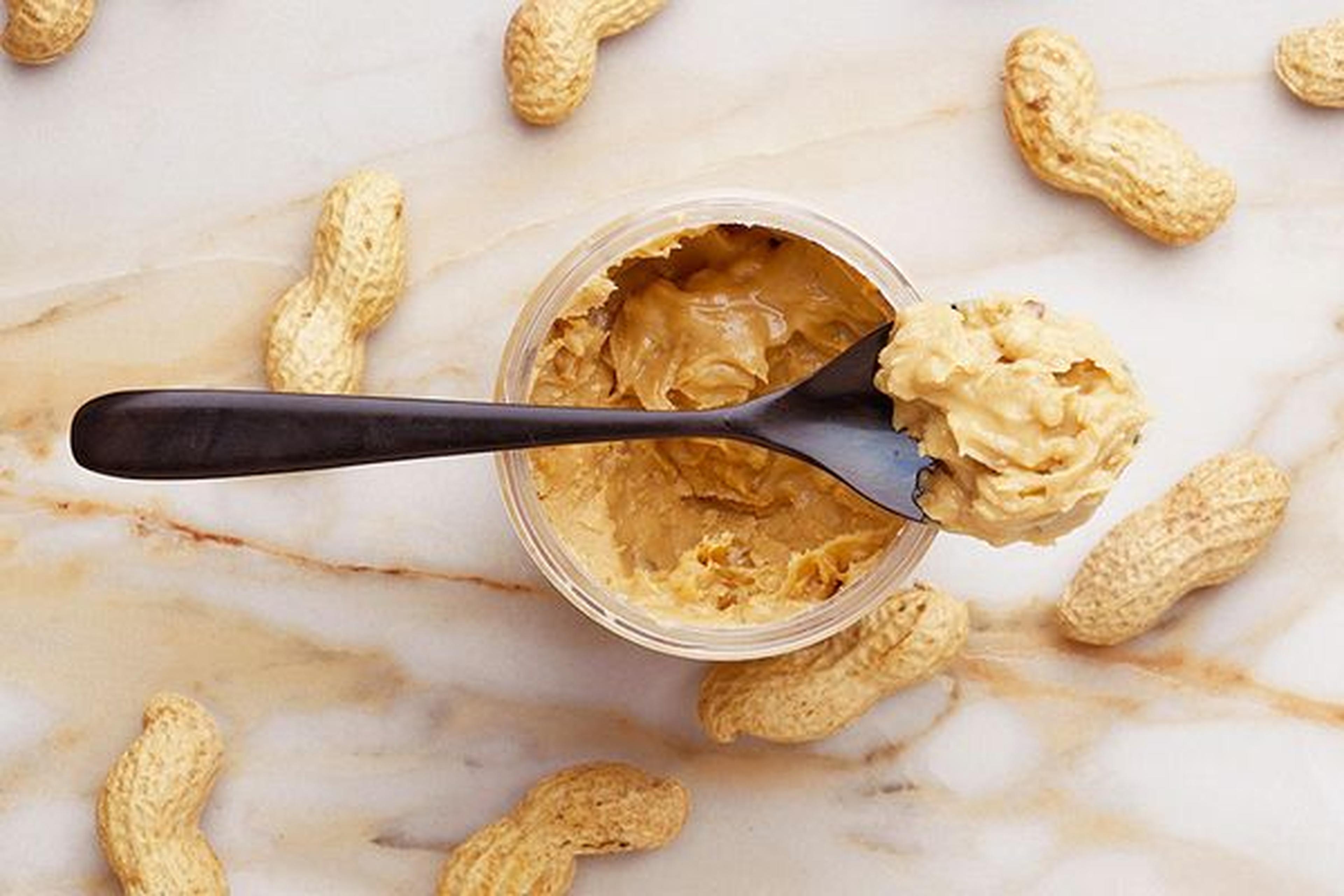 Glas Erdnussbutter mit einem schwarzen Löffel darin, umgeben von ungeschälten Erdnüssen auf einer Marmoroberfläche.