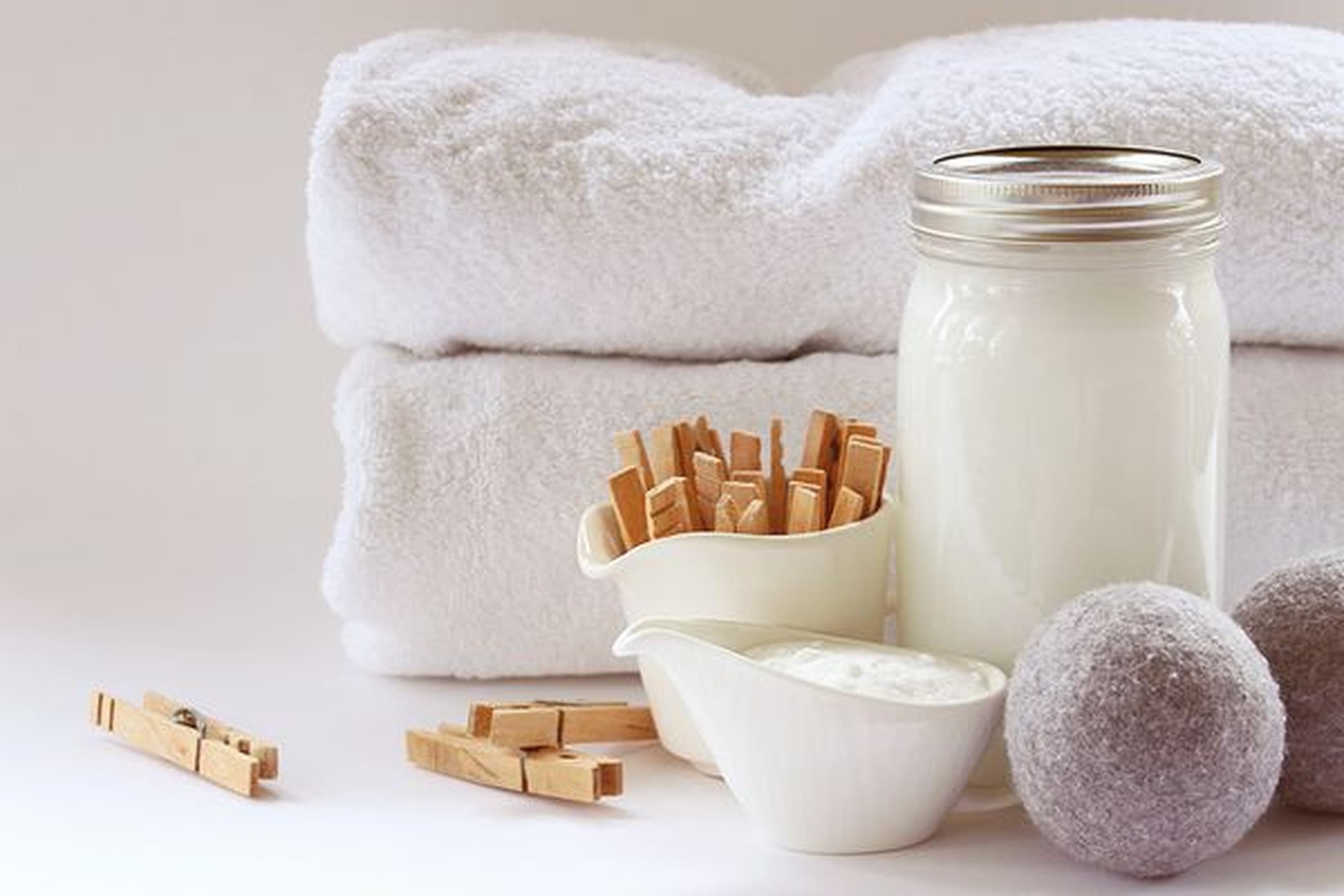 Glas mit weißer Flüssigkeit, kleine Schüssel mit Pulver, Wäscheklammern aus Holz, weiße Handtücher und graue Trocknerbälle aus Wolle auf einer weißen Oberfläche.