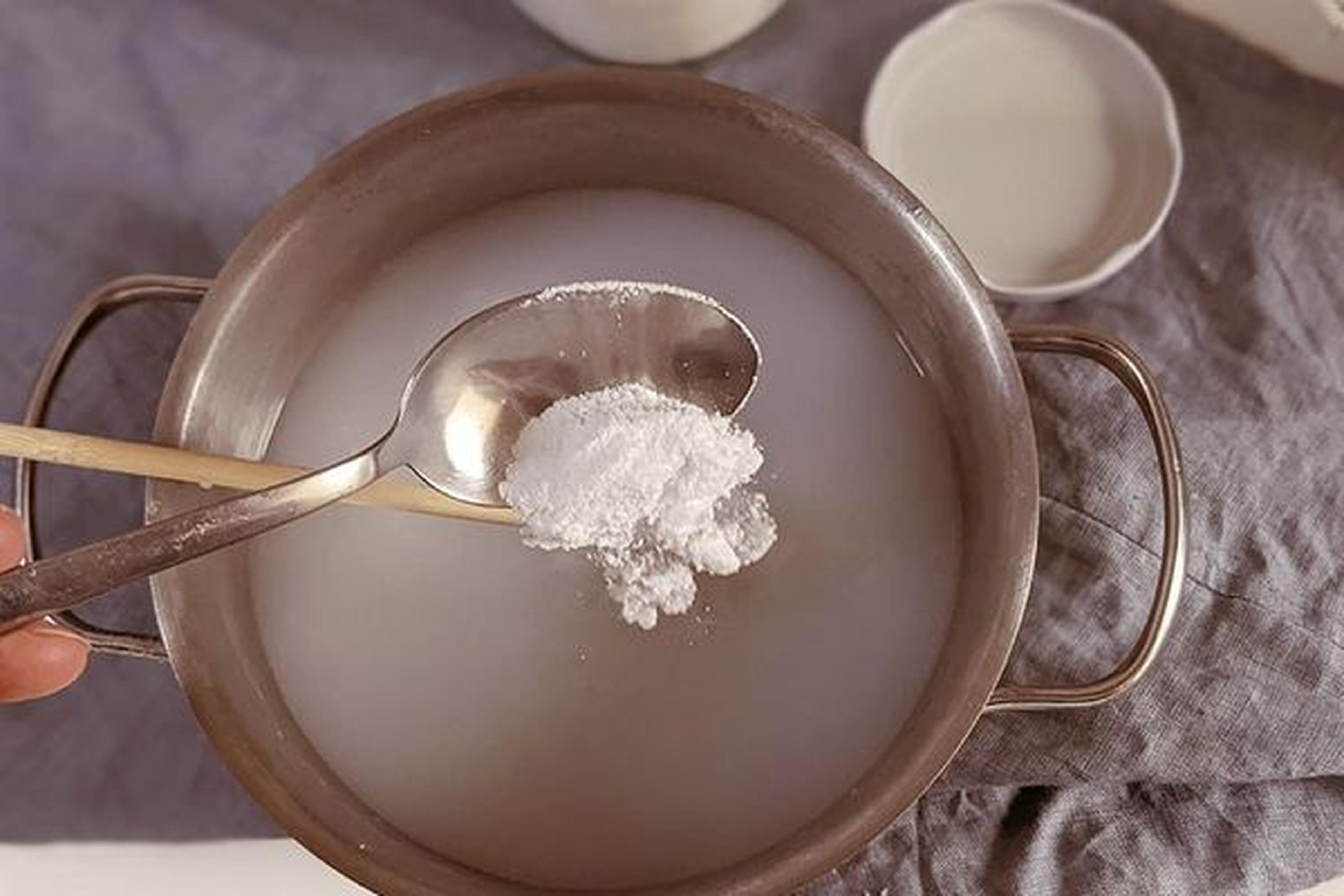 Ein Topf mit Flüssigkeit, über dem ein Löffel mit einer gehäuften Menge weißem Pulver hängt, auf einem grauen Tuch platziert.