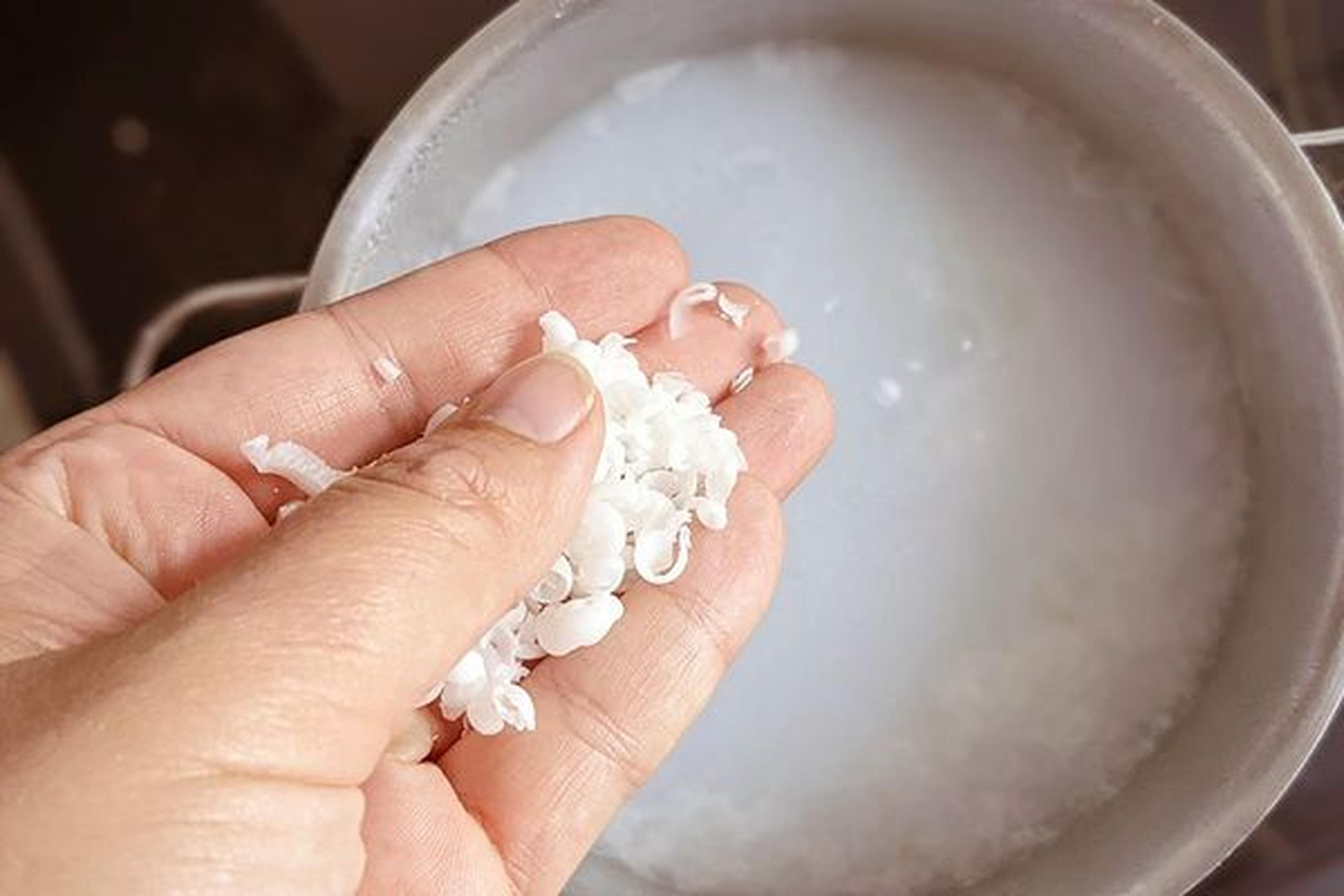 Hand, die Seifenraspeln über einen Topf mit milchiger Flüssigkeit hält, wahrscheinlich bei der Herstellung einer selbstgemachten Seifenlösung.