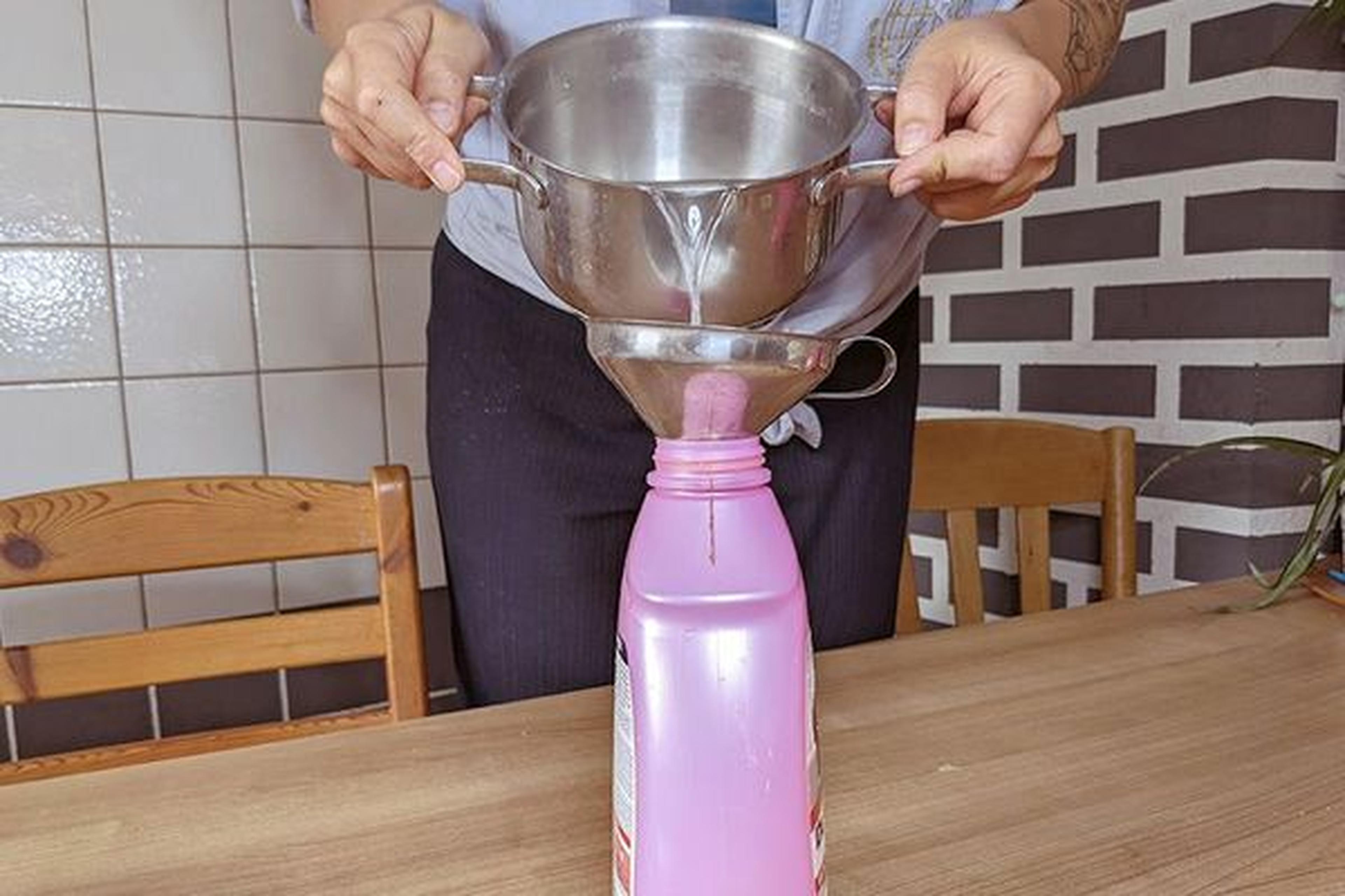 Person, die Flüssigkeit aus einer Metallschüssel durch einen Trichter in eine rosa Plastikflasche auf einem Holztisch gießt, mit gekachelten Wänden im Hintergrund.