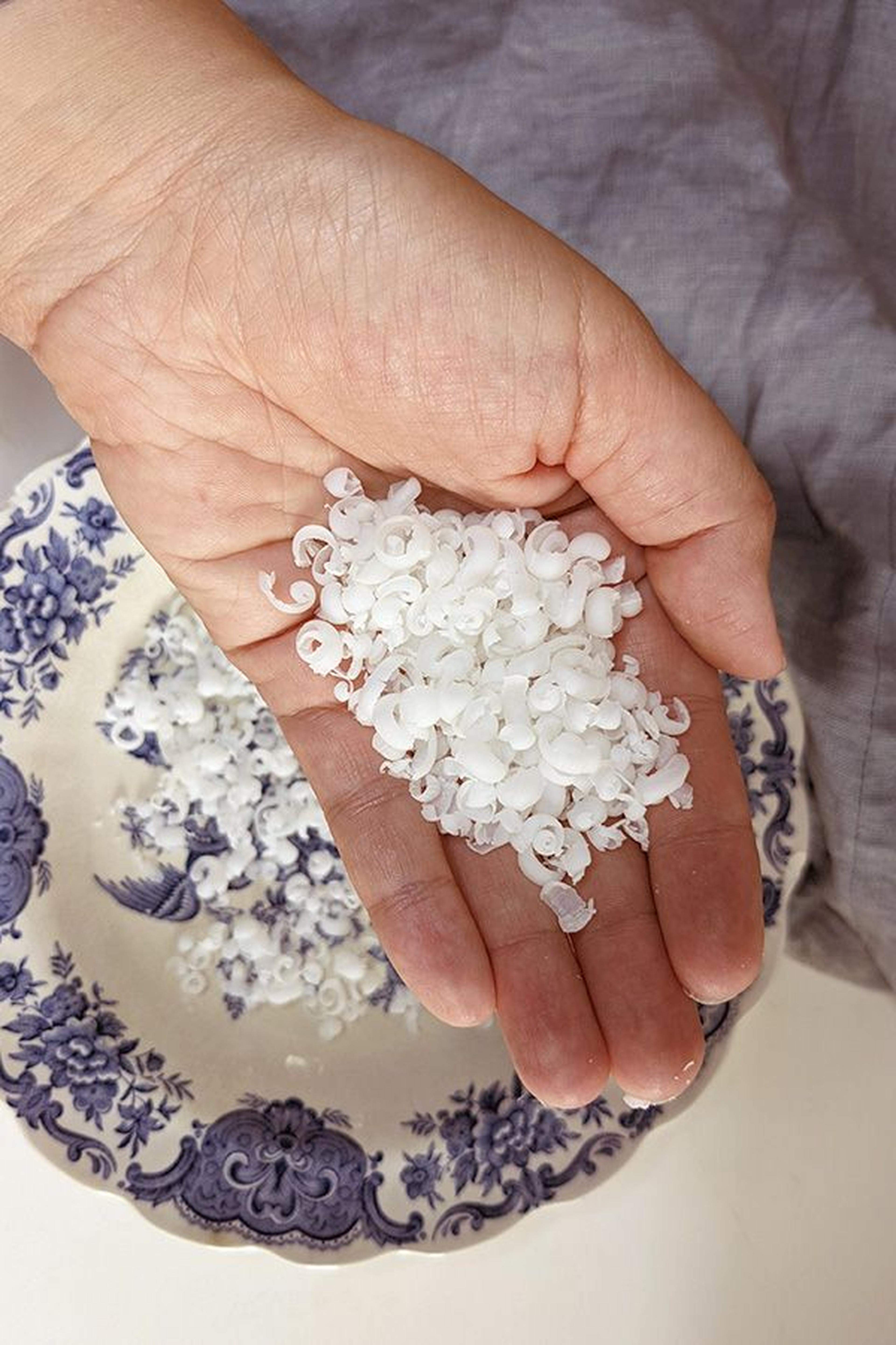 Hand, die kleine weiße Seifenflocken über einem dekorativen Teller mit Blumenmuster hält.