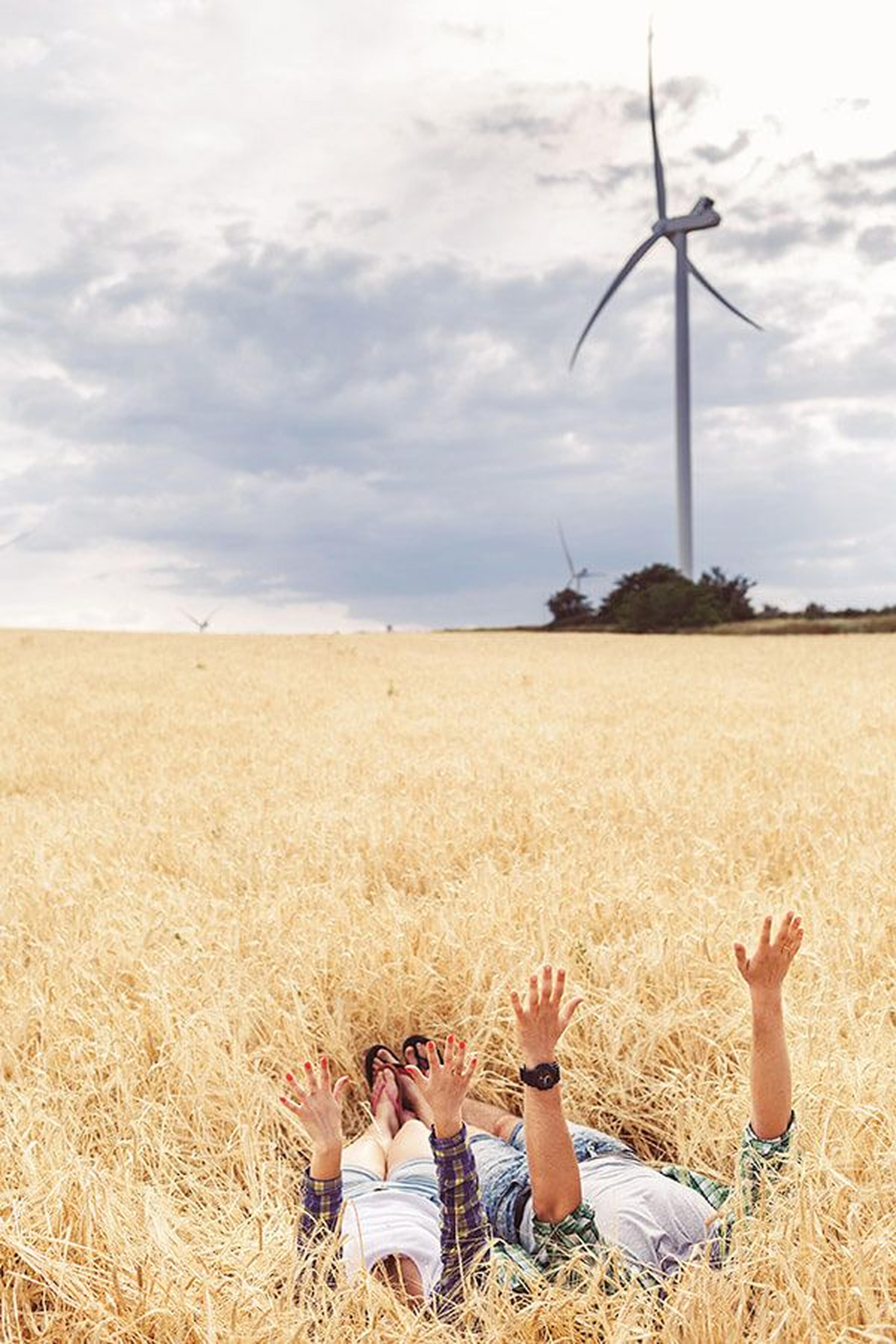 Zwei Menschen liegen mit erhobenen Armen in einem Weizenfeld, im Hintergrund Windräder unter einem bewölkten Himmel.