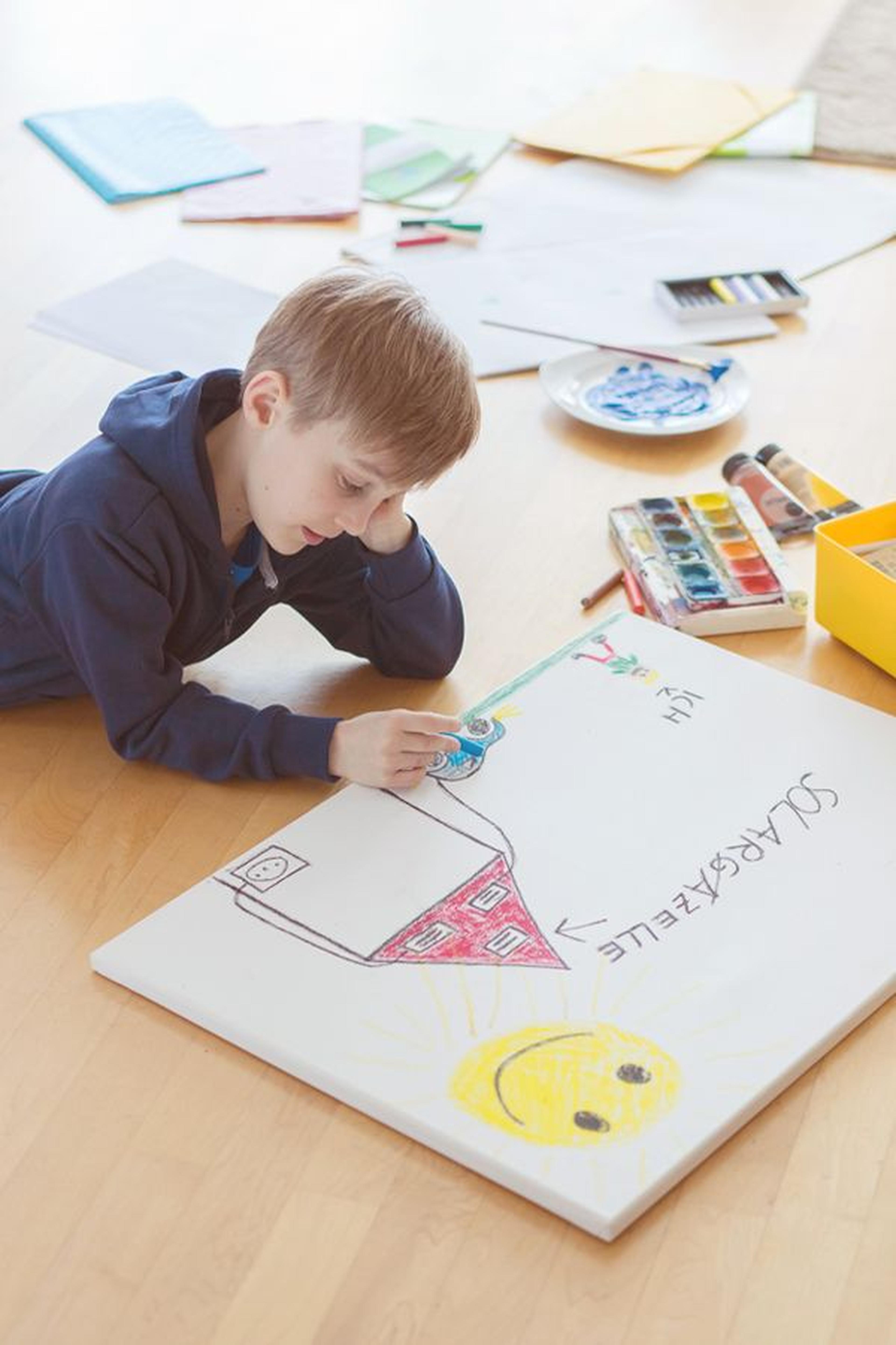 Junge liegt auf dem Boden und malt mit Filzstiften ein buntes Haus und eine Sonne auf eine Leinwand. Malutensilien liegen verstreut auf dem Holzboden.