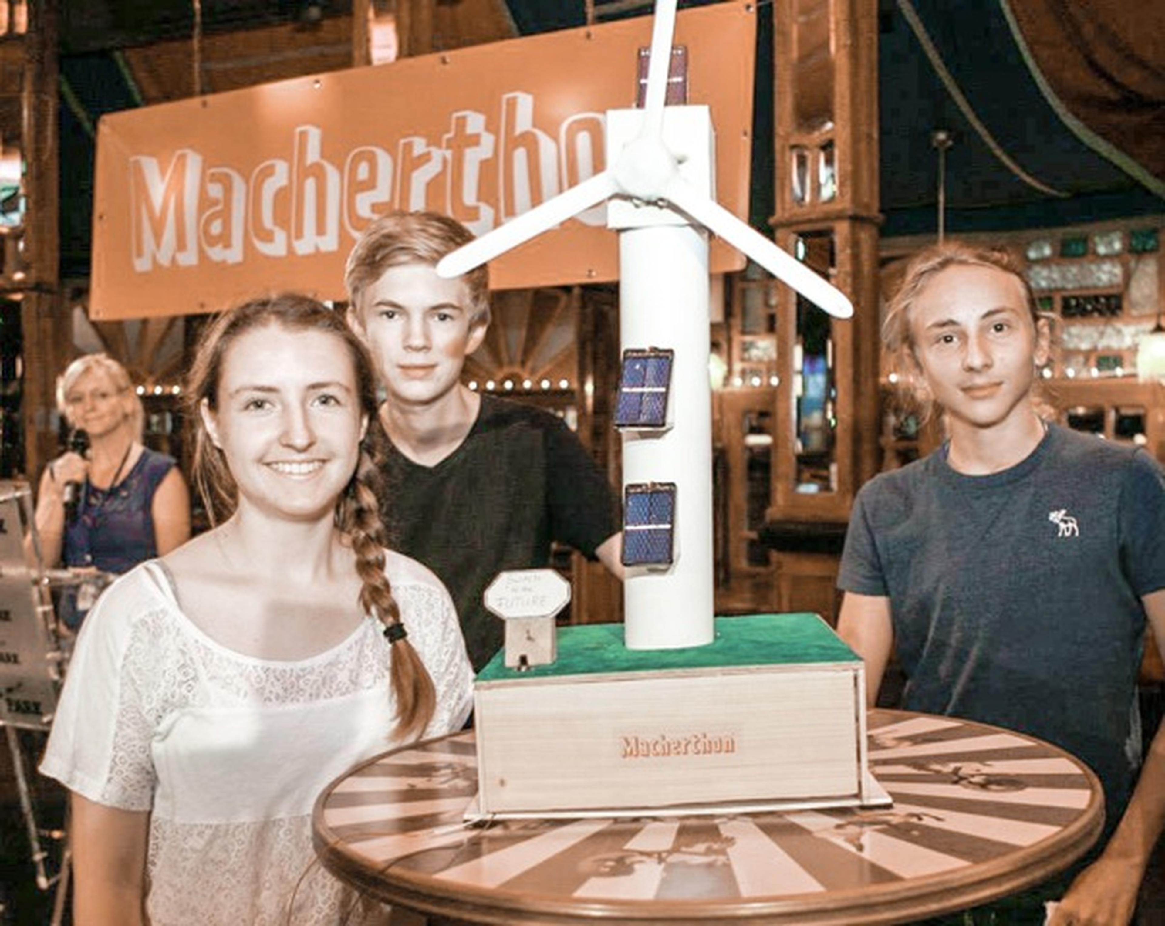 Drei junge Leute stehen um ein Modell einer Windkraftanlage mit Sonnenkollektoren auf einem Tisch mit der Aufschrift „Macherthon“.