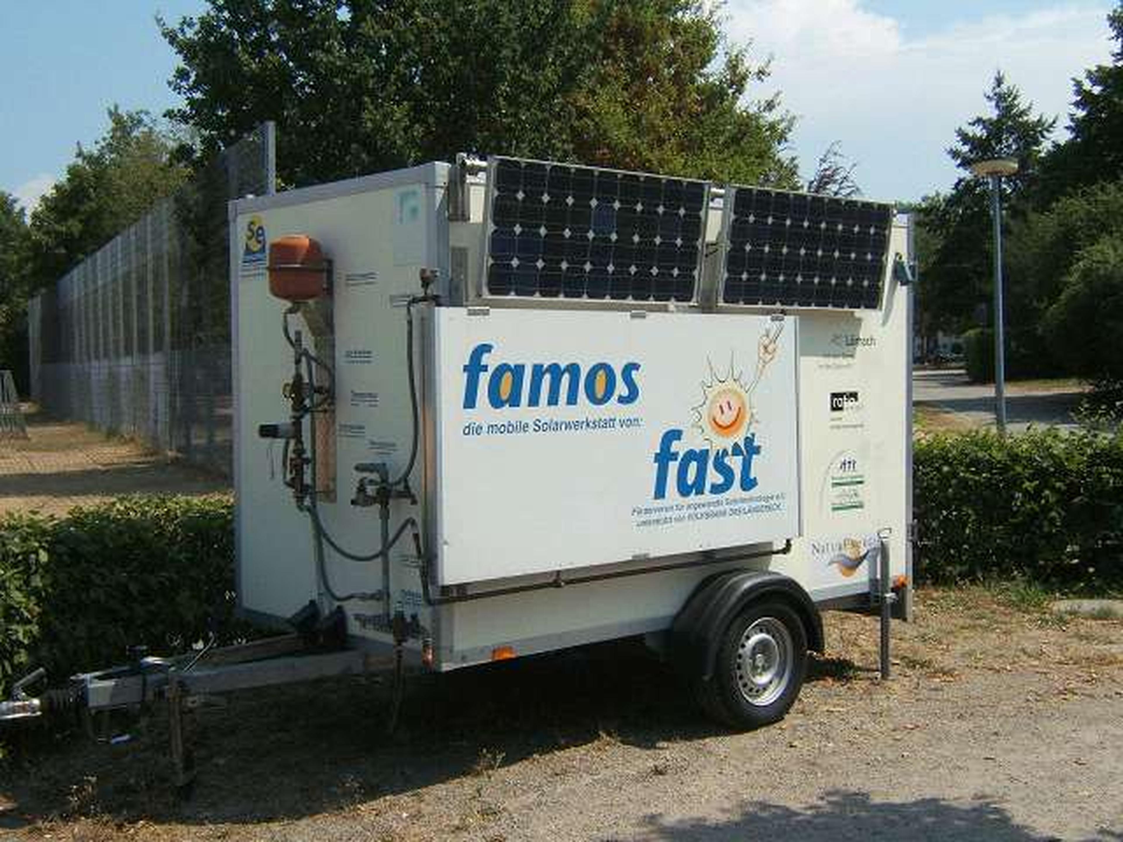 Ein mobiler Werkstattanhänger mit Sonnenkollektoren, an dessen Seite die Aufschriften „famos“ und „fast“ zu sehen sind, steht im Freien in der Nähe von Grünflächen.