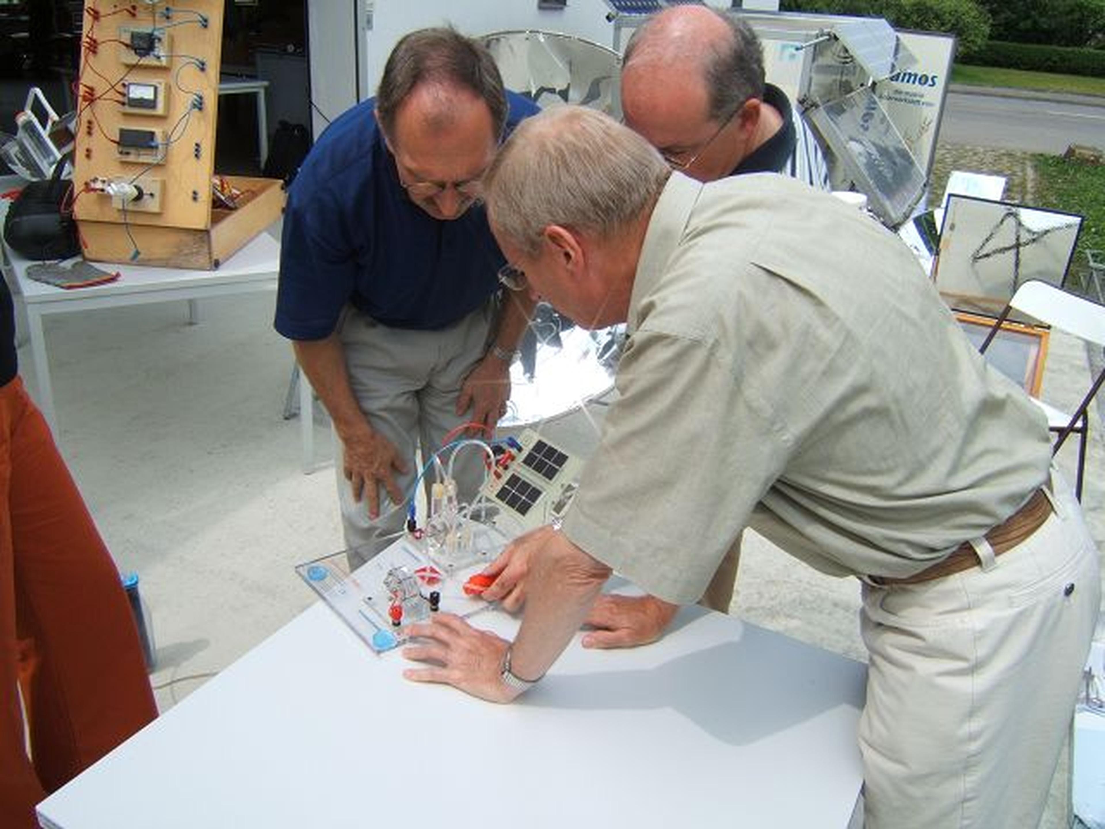 An einem sonnigen Tag untersuchen drei Männer im Freien auf einem weißen Tisch ein solarbetriebenes Gerät, umgeben von technischer Ausrüstung und Displays.