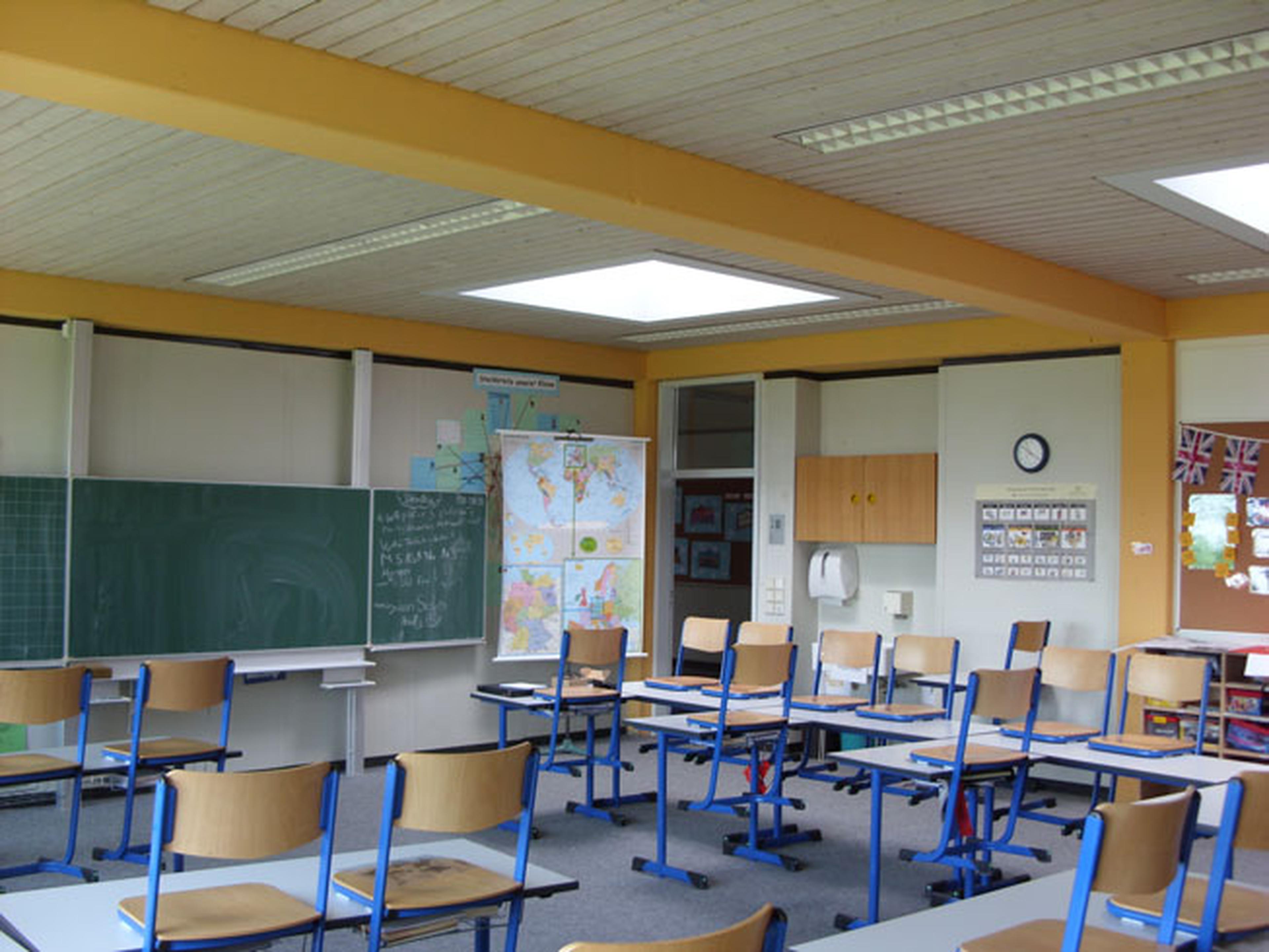 Ein leeres Klassenzimmer mit Tischen, Stühlen, einer Tafel mit Schriftzügen und Landkarten an der Wand. Man sieht eine helle Beleuchtung und eine Uhr.