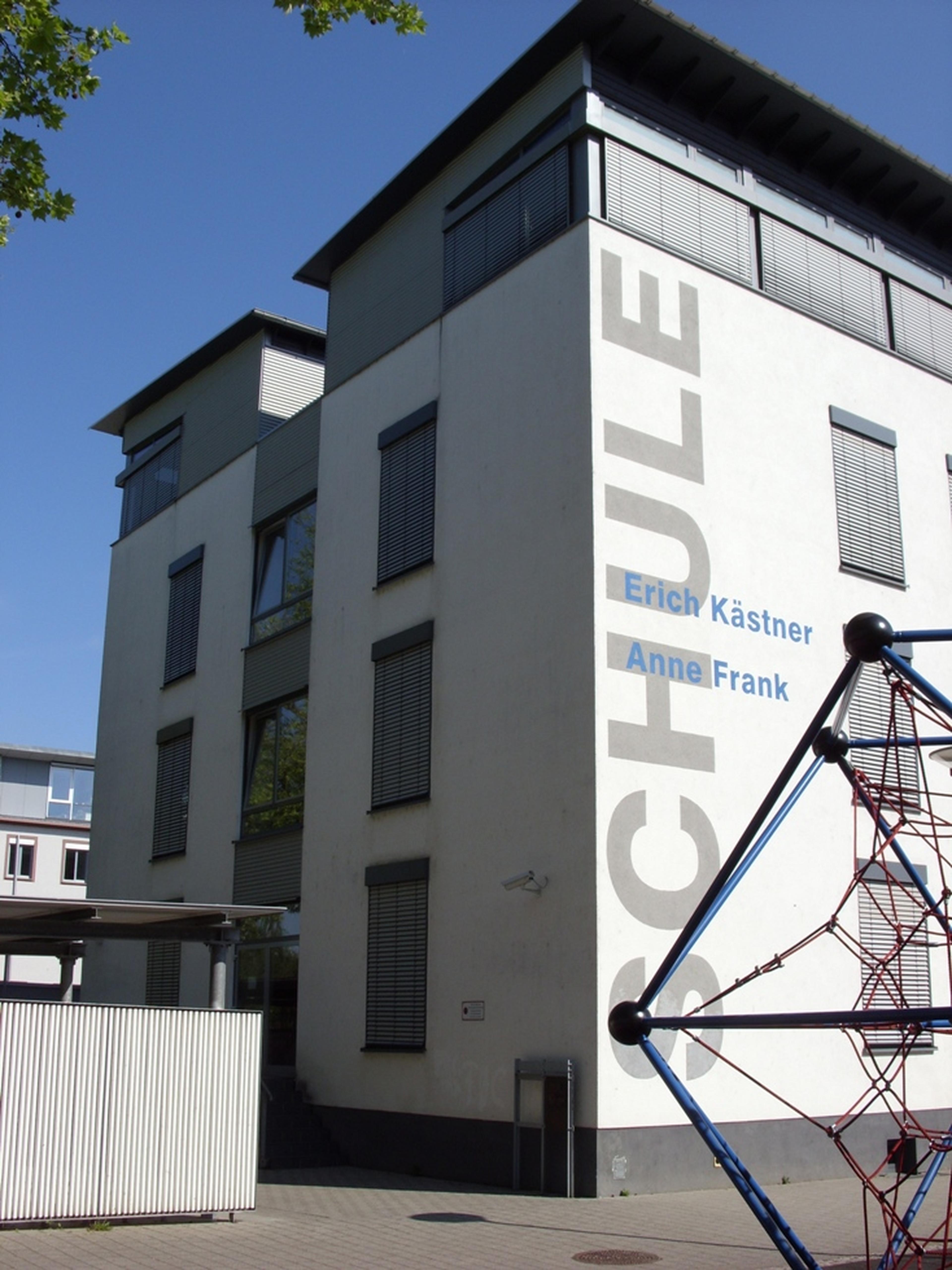 Ein modernes Schulgebäude, an dessen Seite die Namen „Erich Kästner“ und „Anne Frank“ zu lesen sind. Im Vordergrund ist eine Kletteranlage zu sehen.