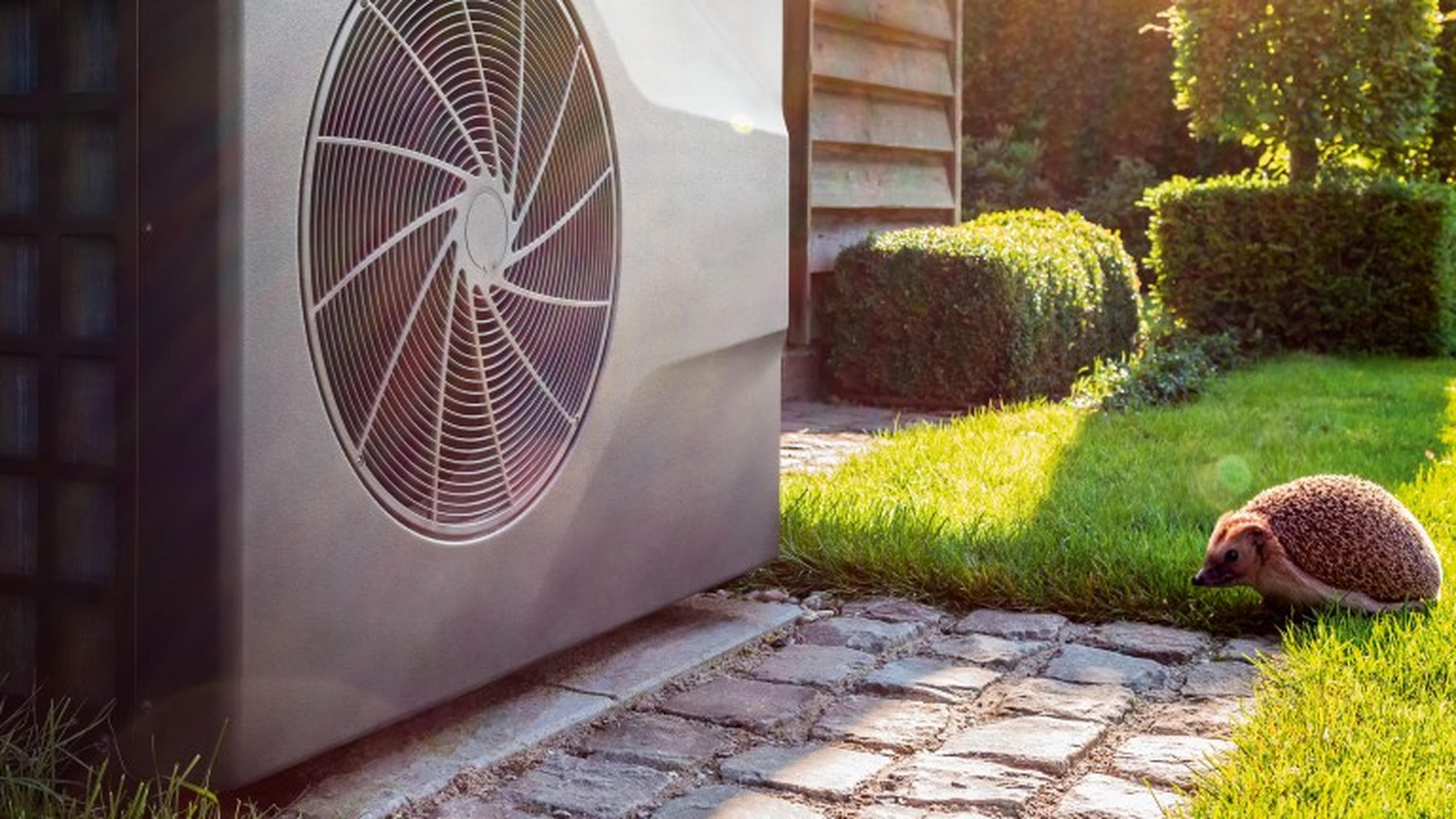 Eine Wärmepumpe im Garten neben einem Haus, auf dem Rasen läuft ein Igel vorbeii.