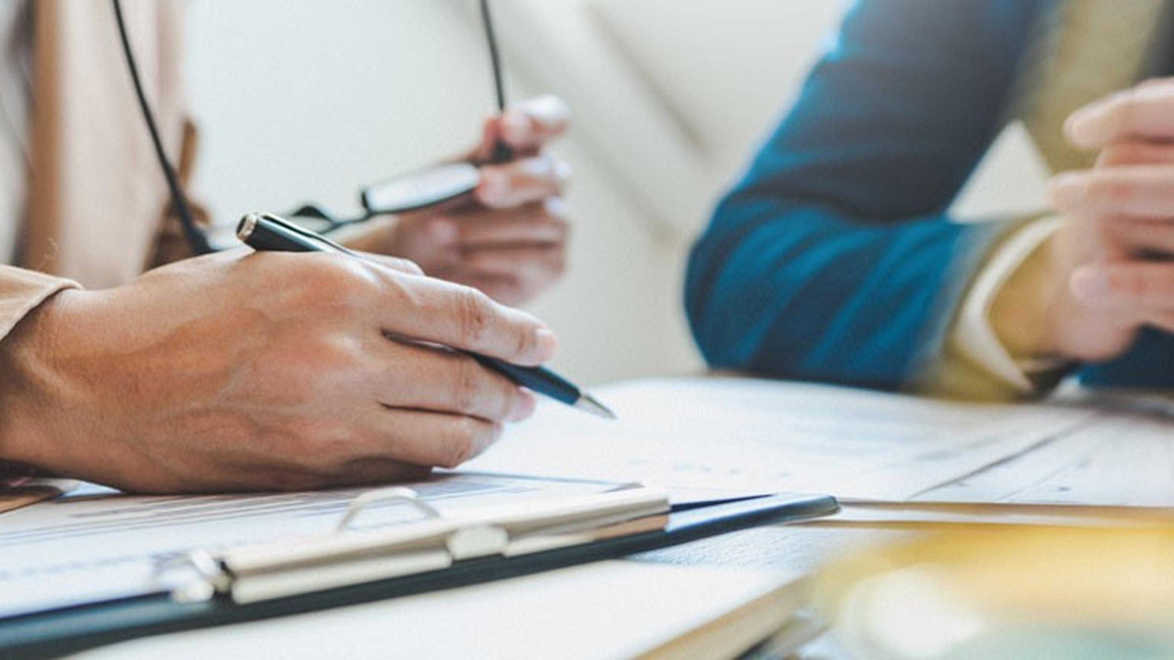 Zwei Personen prüfen Dokumente an einem Tisch und halten Stifte in der Hand. Eine trägt eine beige Jacke, die andere eine blaue. Im Vordergrund ist ein Klemmbrett zu sehen.