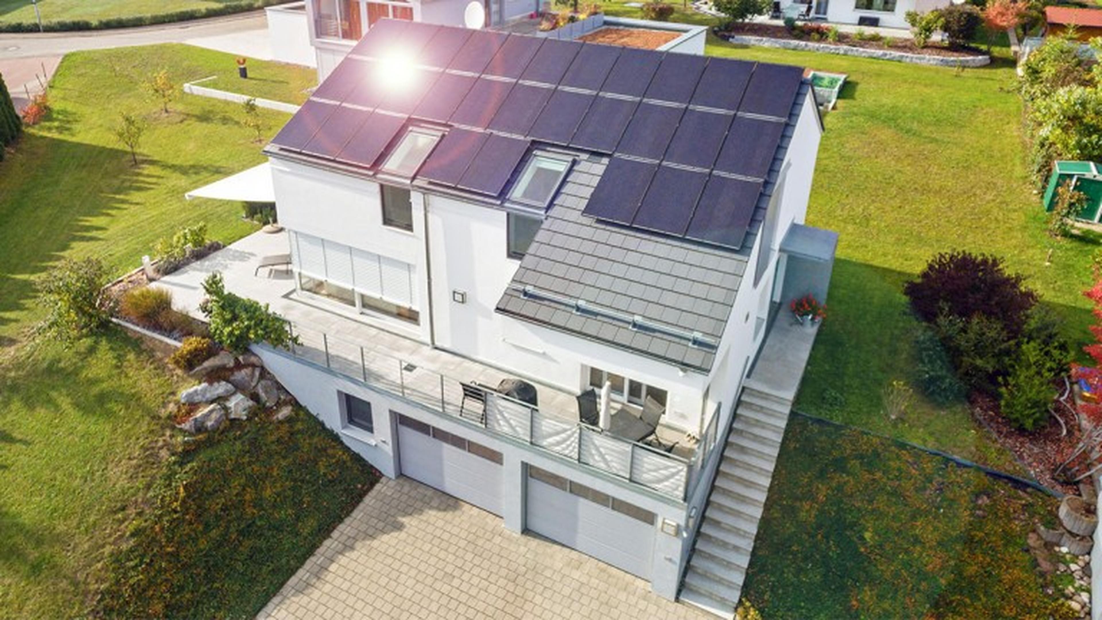 Modernes weißes Haus mit Photovoltaik Paneelen auf dem Dach, umgeben von einem üppig grünen Rasen und einer gepflasterten Auffahrt in einem Wohngebiet.