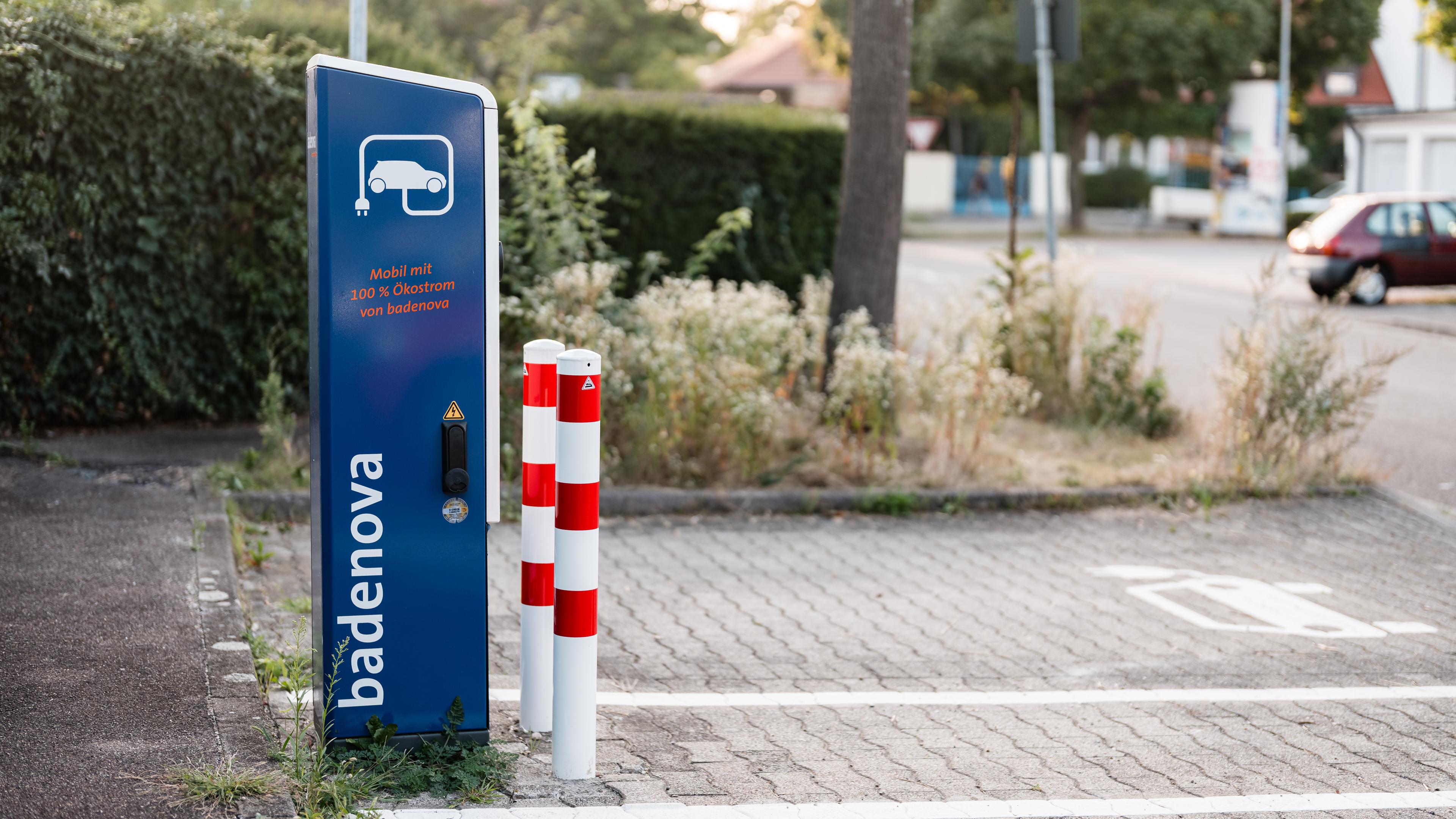 Seitenansicht einer Ladesäule mit zwei weiß-rot-gestreiften Pollern davor und einem Parkplatz dahinter, der mit einem E-Auto-Symbol markiert ist.