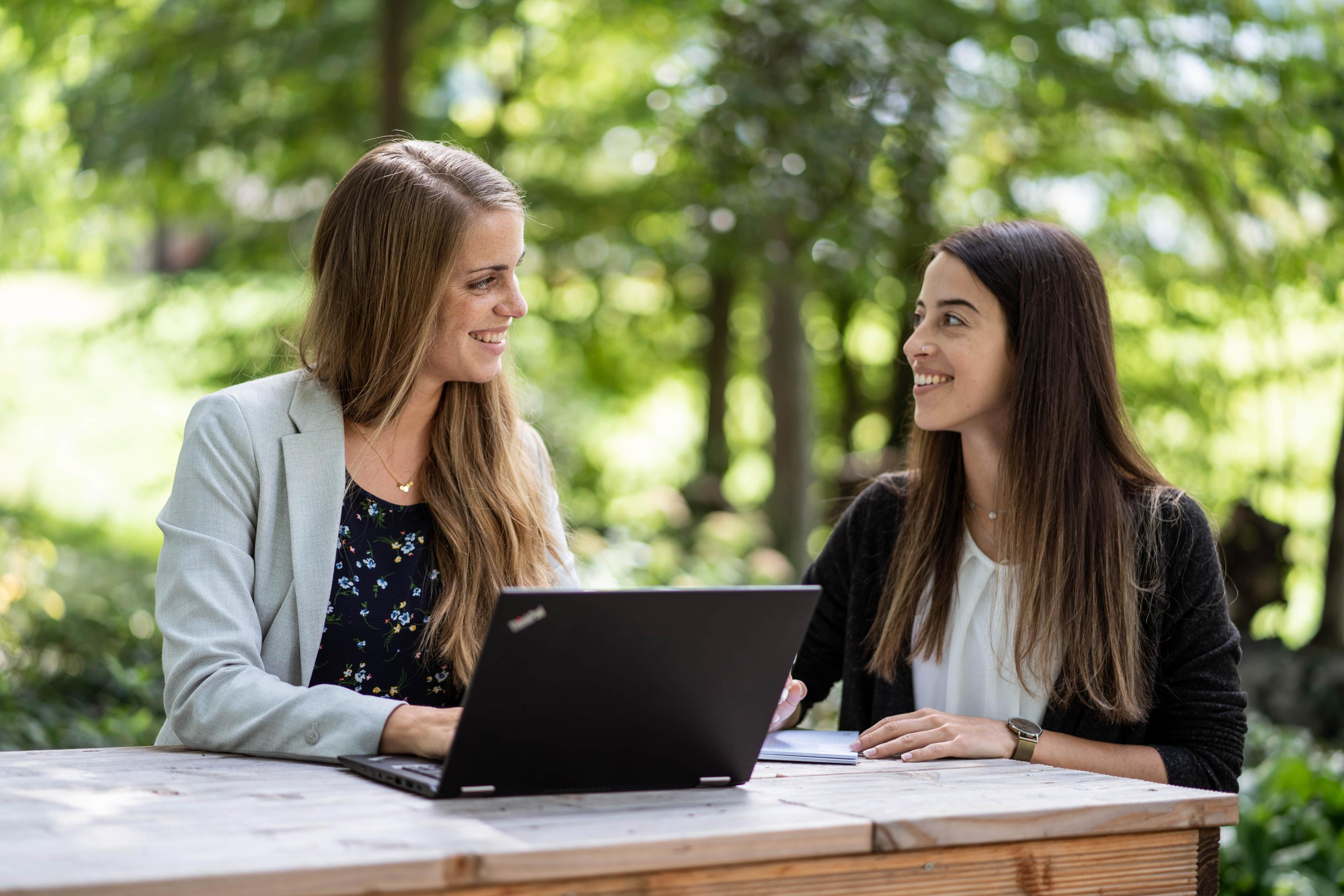 Zwei Frauen sitzen an einem Tisch im Freien mit einem Laptop, lächeln und unterhalten sich, umgeben von viel Grün.