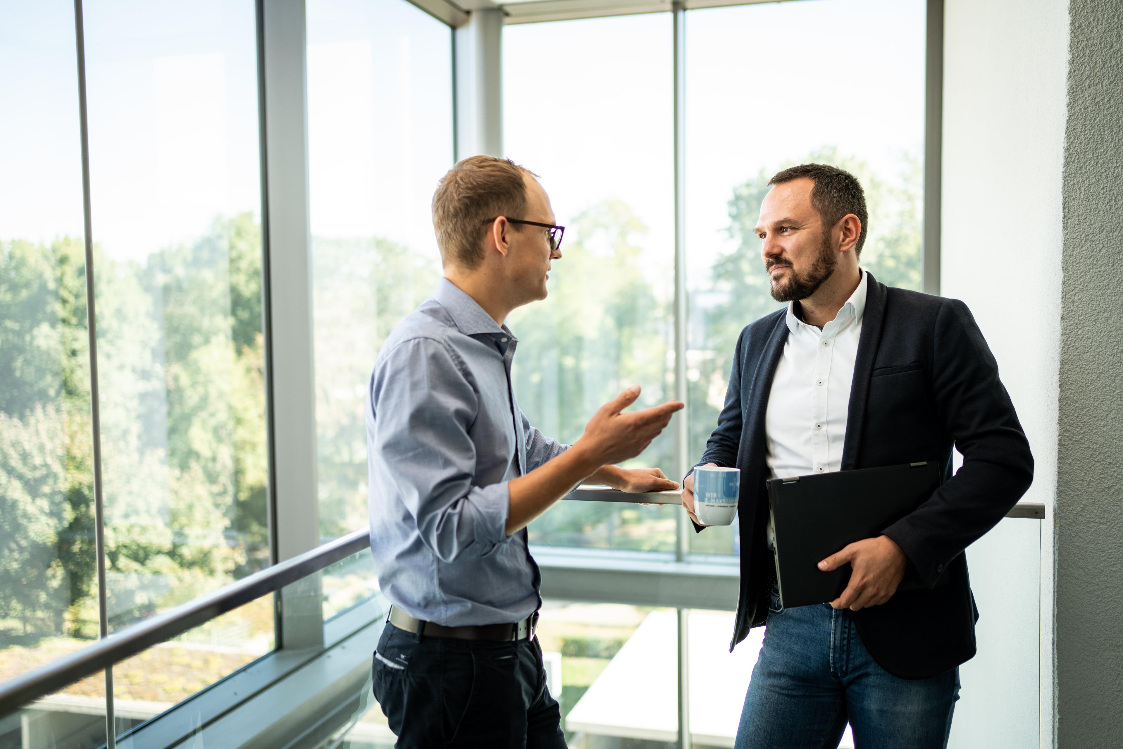 Zwei Männer in einem modernen Büro, einer mit einer Tasse und einer Mappe in der Hand, unterhalten sich in der Nähe großer Fenster mit Blick auf die grüne Umgebung.