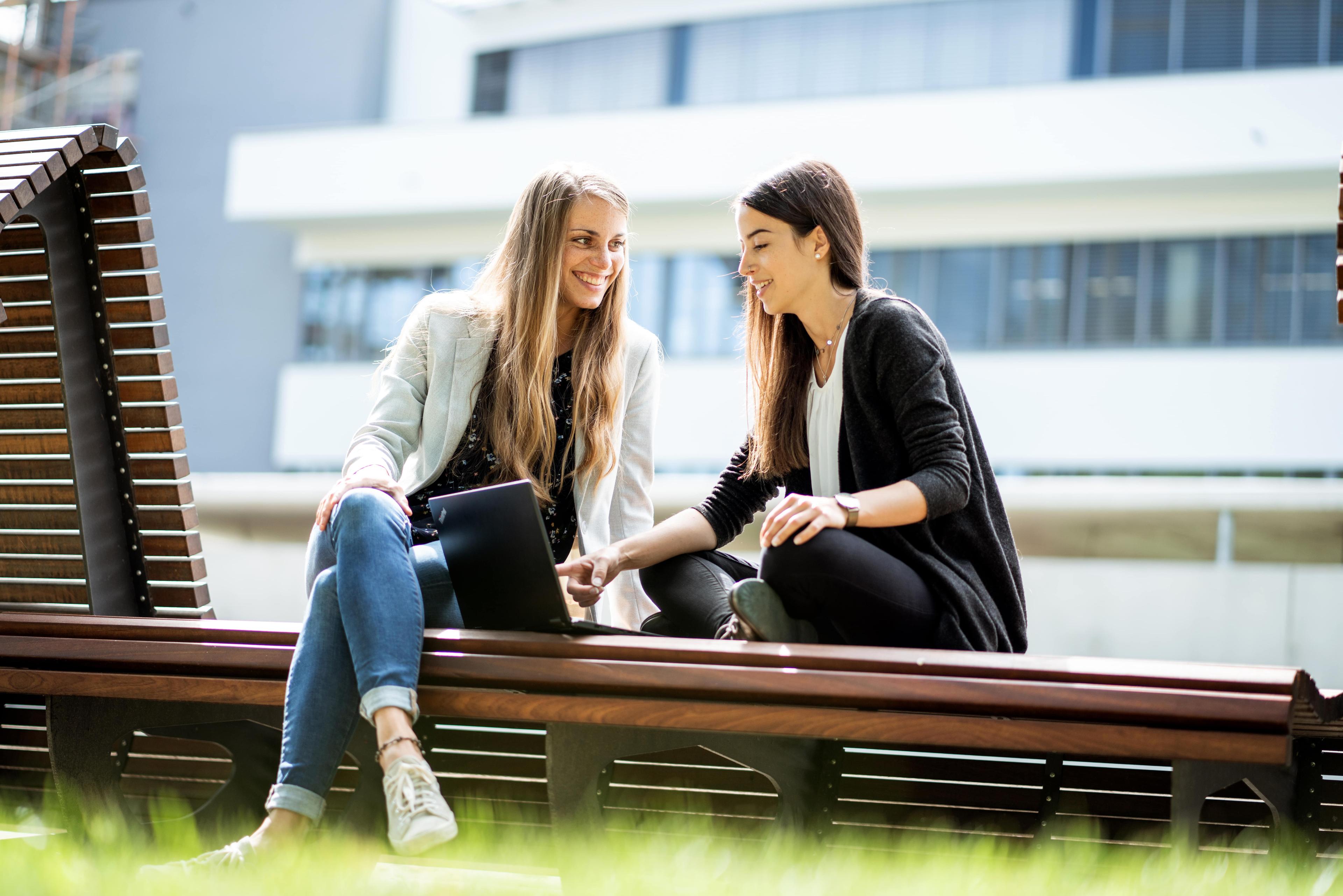 Zwei Frauen sitzen auf einer Bank im Freien, lächeln und schauen auf einen Laptop, im Hintergrund ist ein modernes Gebäude zu sehen.
