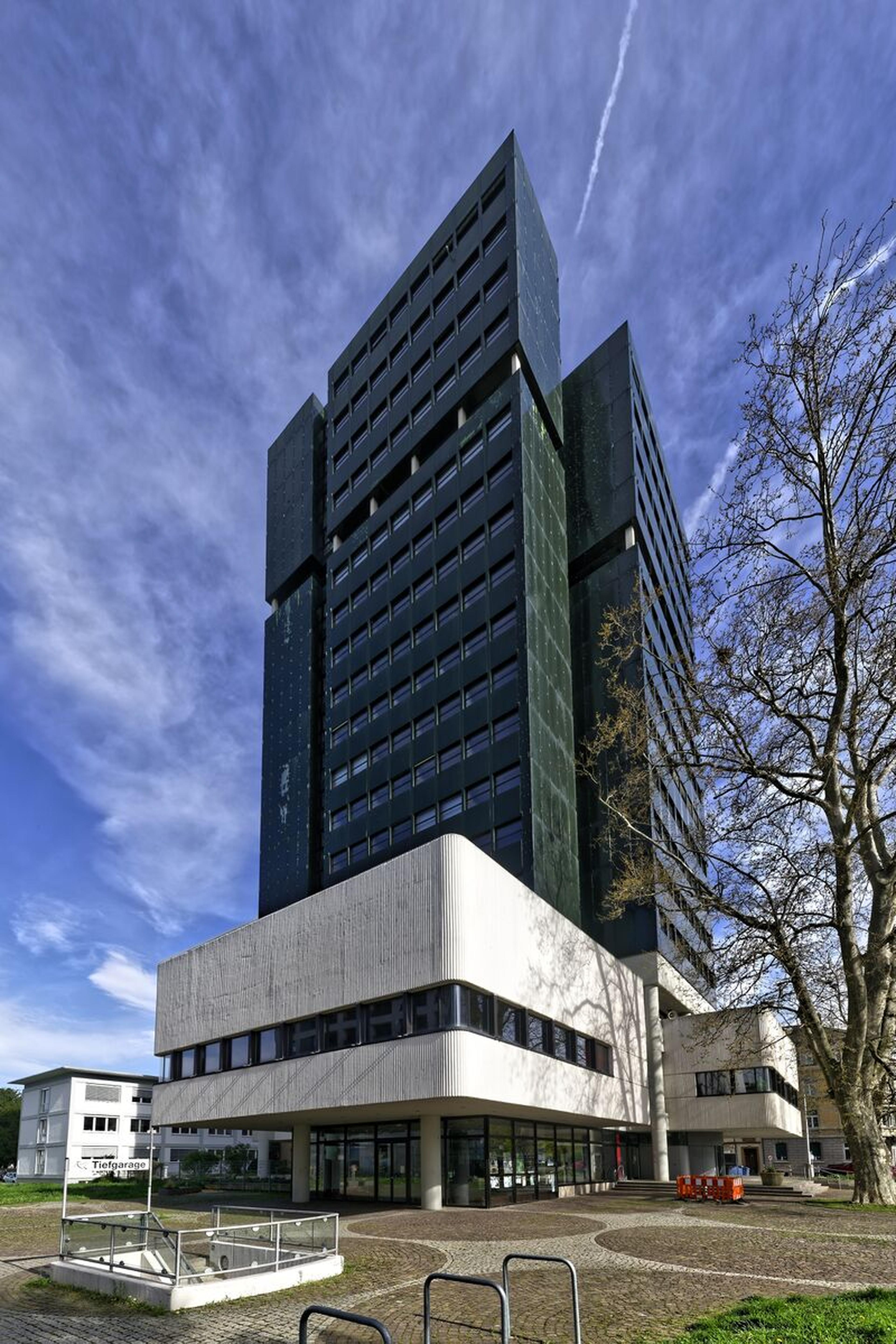 Ein hohes, modernes Gebäude mit dunkler Fassade und weißem Sockel vor blauem Himmel mit vereinzelten Wolken und kahlen Bäumen in der Nähe.