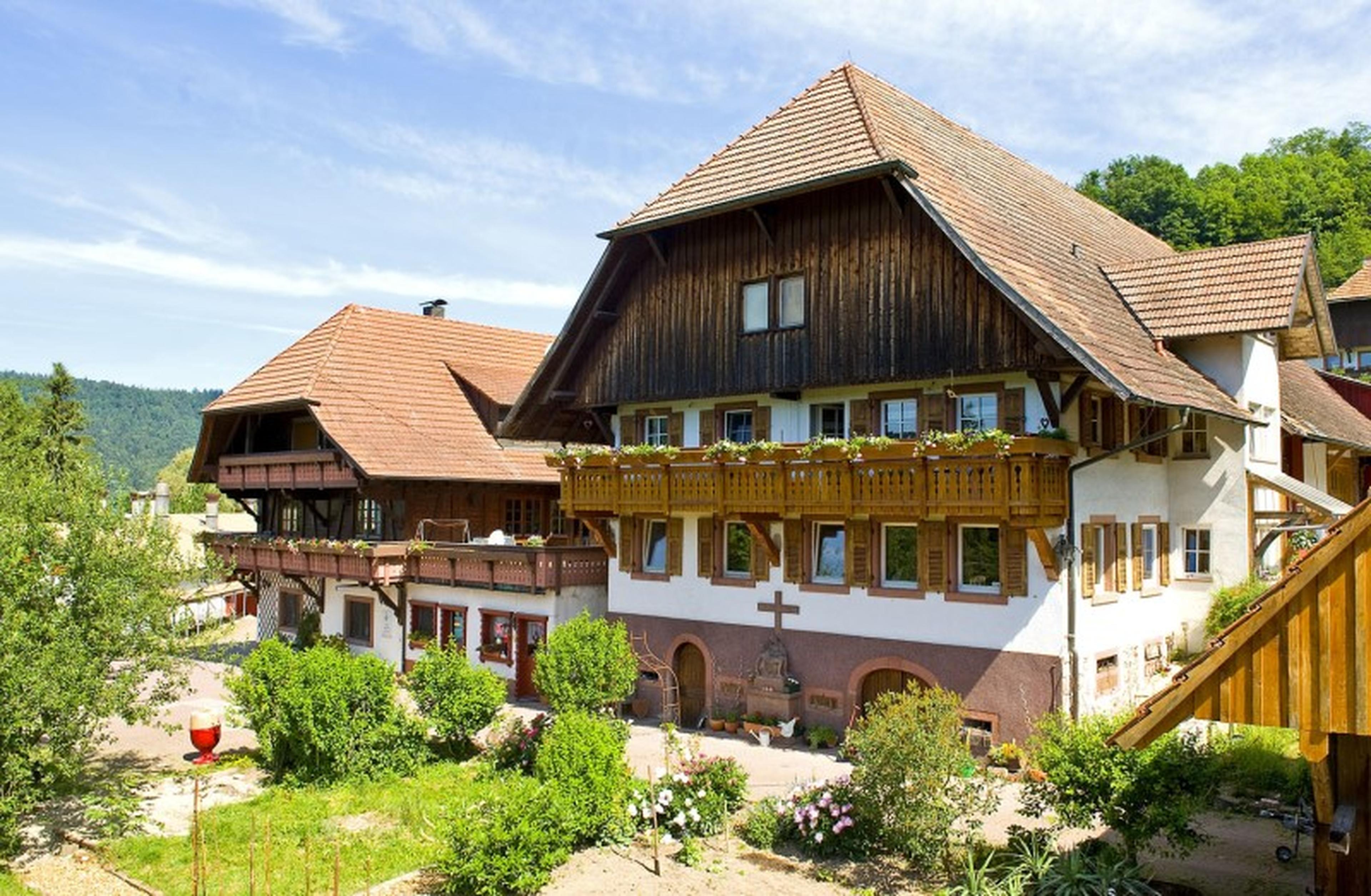 Ein traditionelles deutsches Bauernhaus mit Holzbalkonen und rotem Ziegeldach, umgeben von viel Grün unter strahlend blauem Himmel.