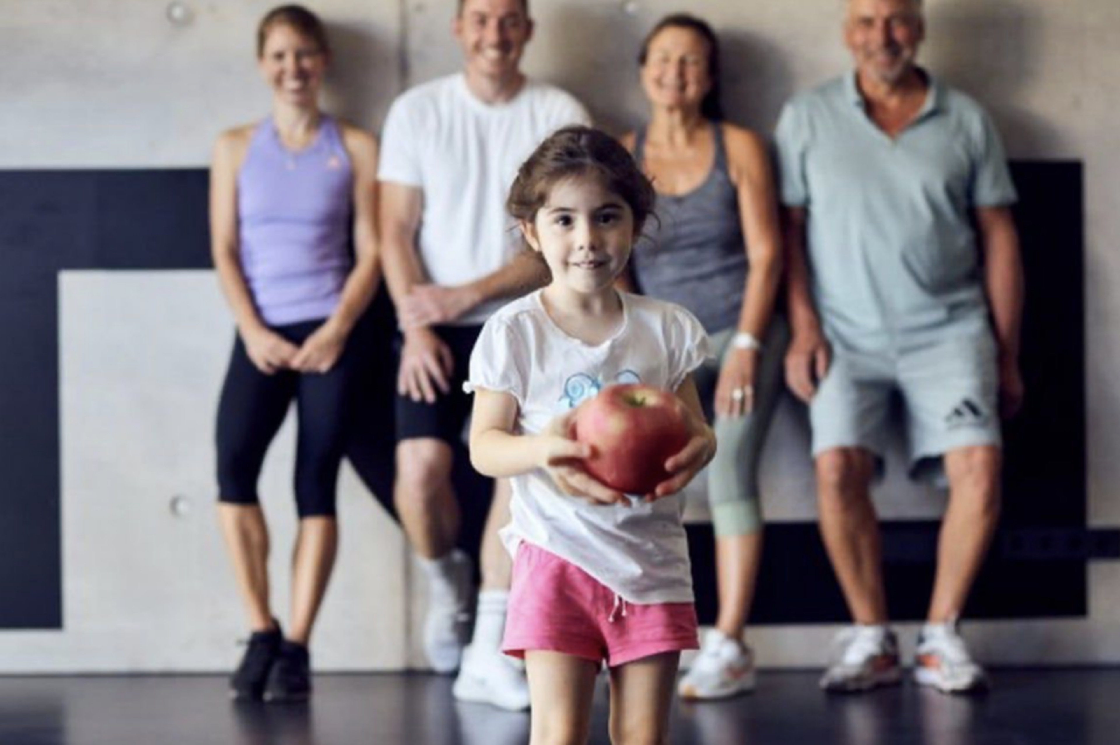 Ein junges Mädchen, das einen roten Ball in der Hand hält, steht vor vier Erwachsenen, die in einer Turnhalle an einer Wand lehnen.