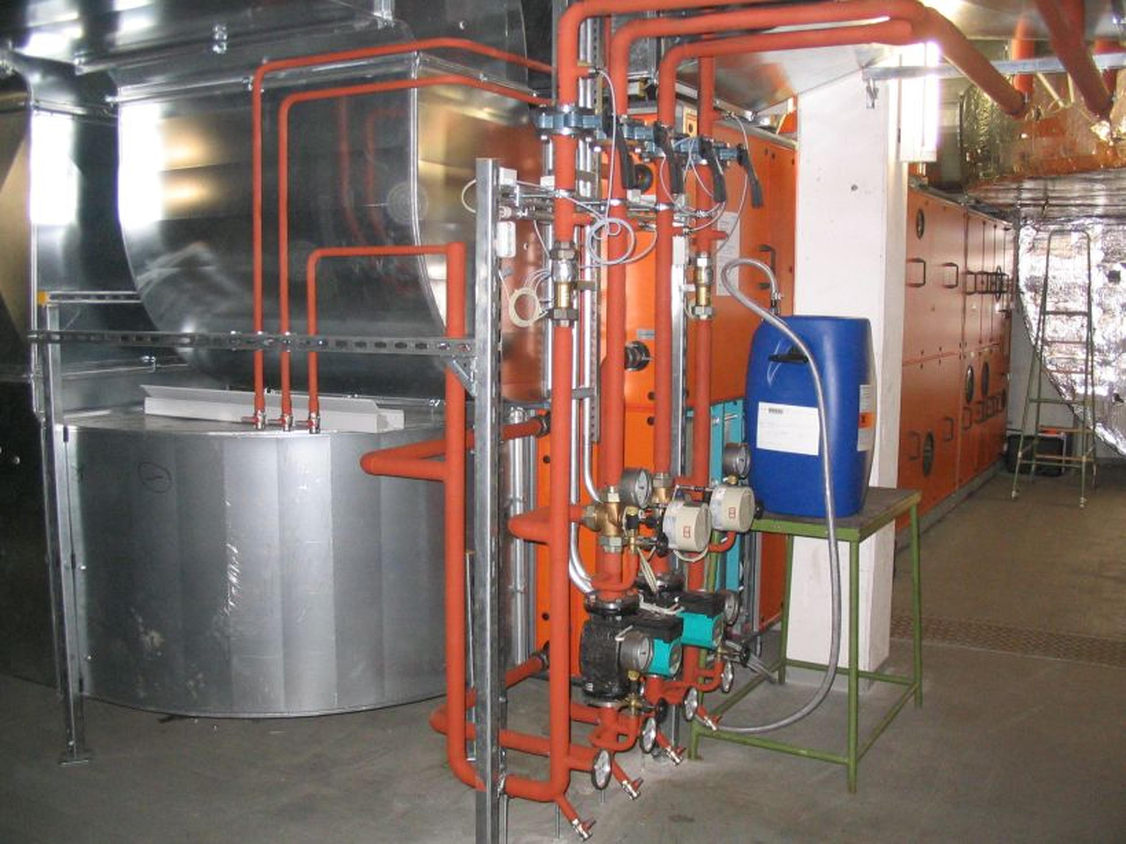 Industrielle Heizungsanlage mit roten Rohren, Ventilen und einem blauen Behälter. Im Hintergrund sind Metalltanks und Maschinen zu sehen.