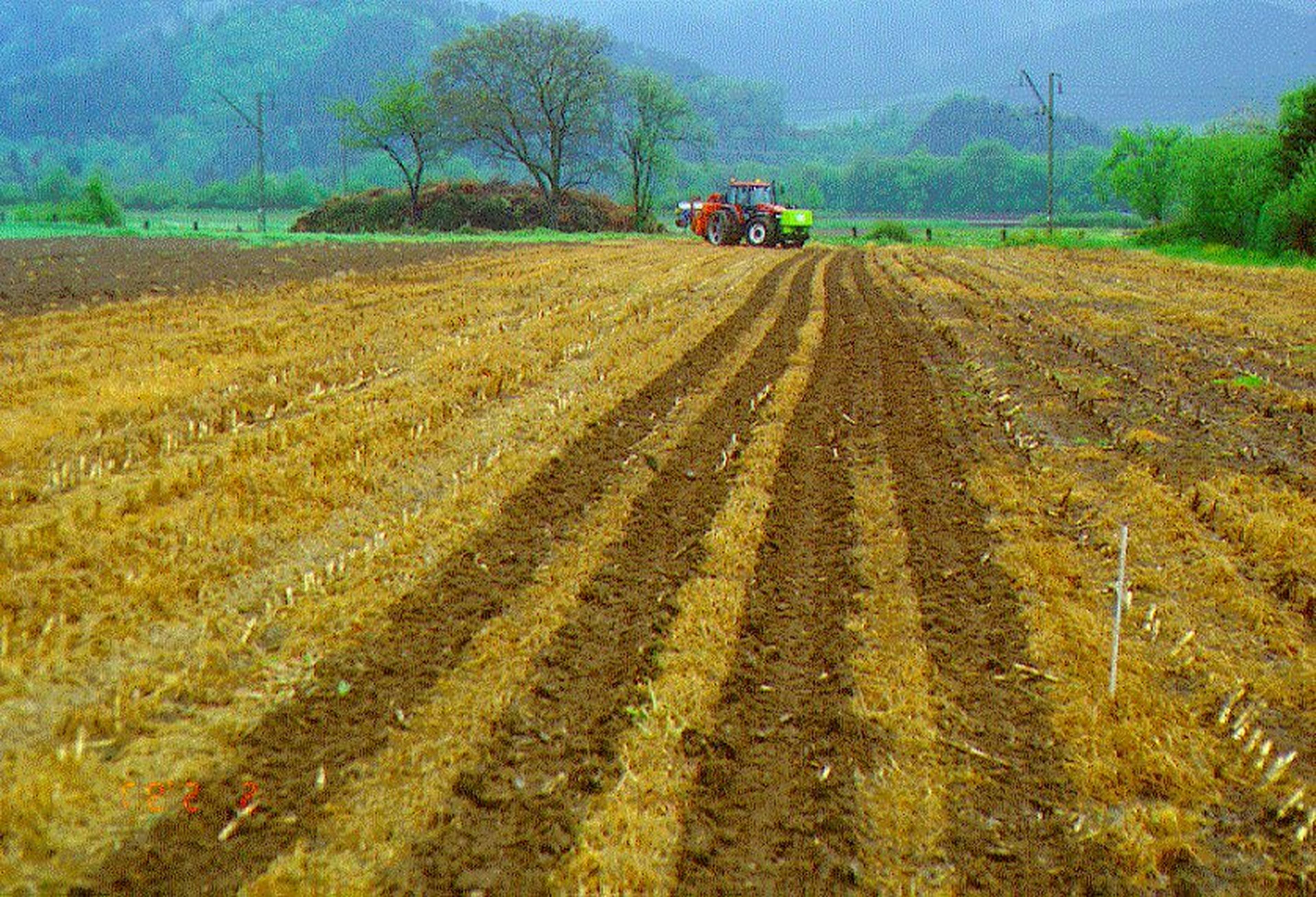 Ein Traktor pflügt ein Feld in einer ländlichen Landschaft mit Bäumen in der Ferne und einem dunstigen Bergpanorama im Hintergrund.