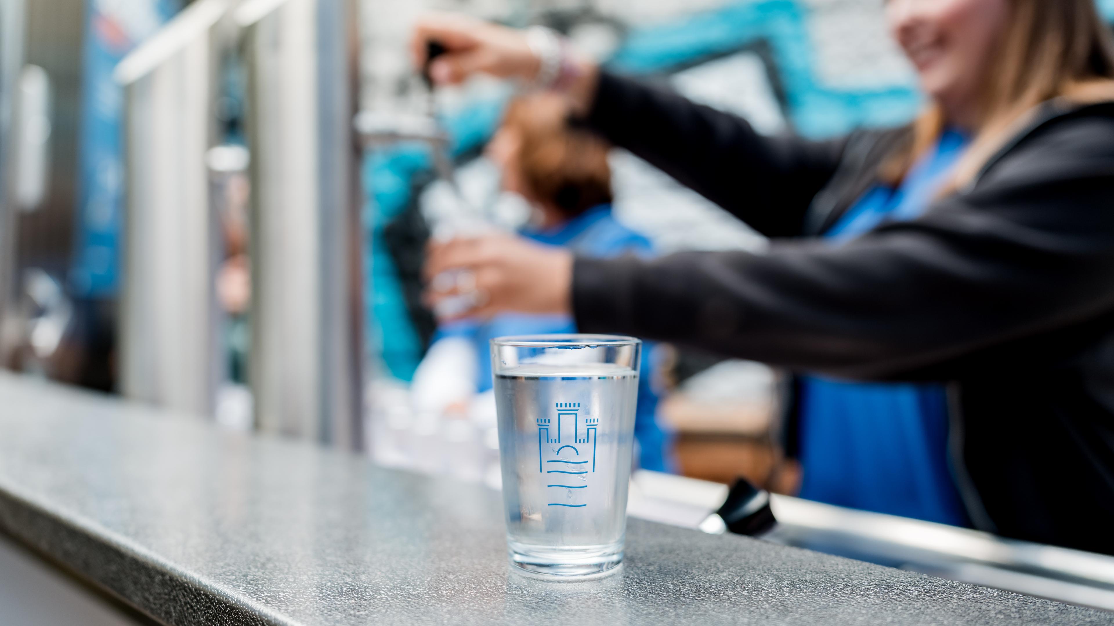 Auf einer Theke steht ein Glas Wasser, im Hintergrund sind Menschen in blauen Hemden zu sehen, die in der Nähe eines Wasserspenders verschwommen wirken.