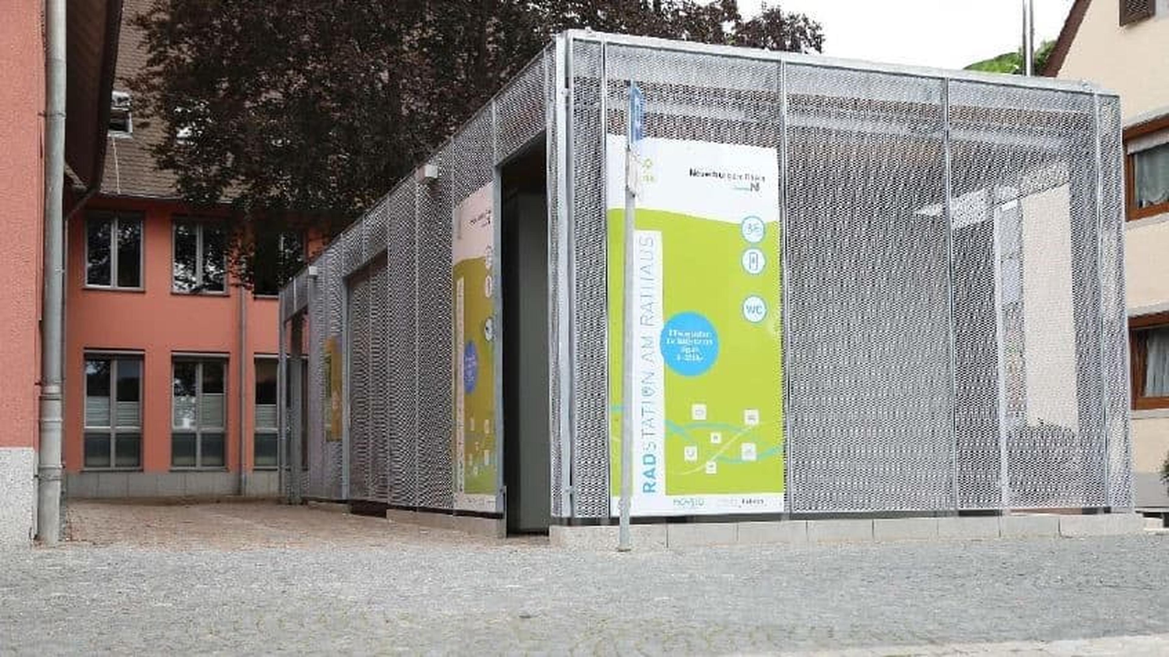 Moderne Fahrradabstellanlage mit Metallgitterwänden und einem farbenfrohen Informationsbanner. Befindet sich zwischen Gebäuden auf einem Kopfsteinpflasterweg.