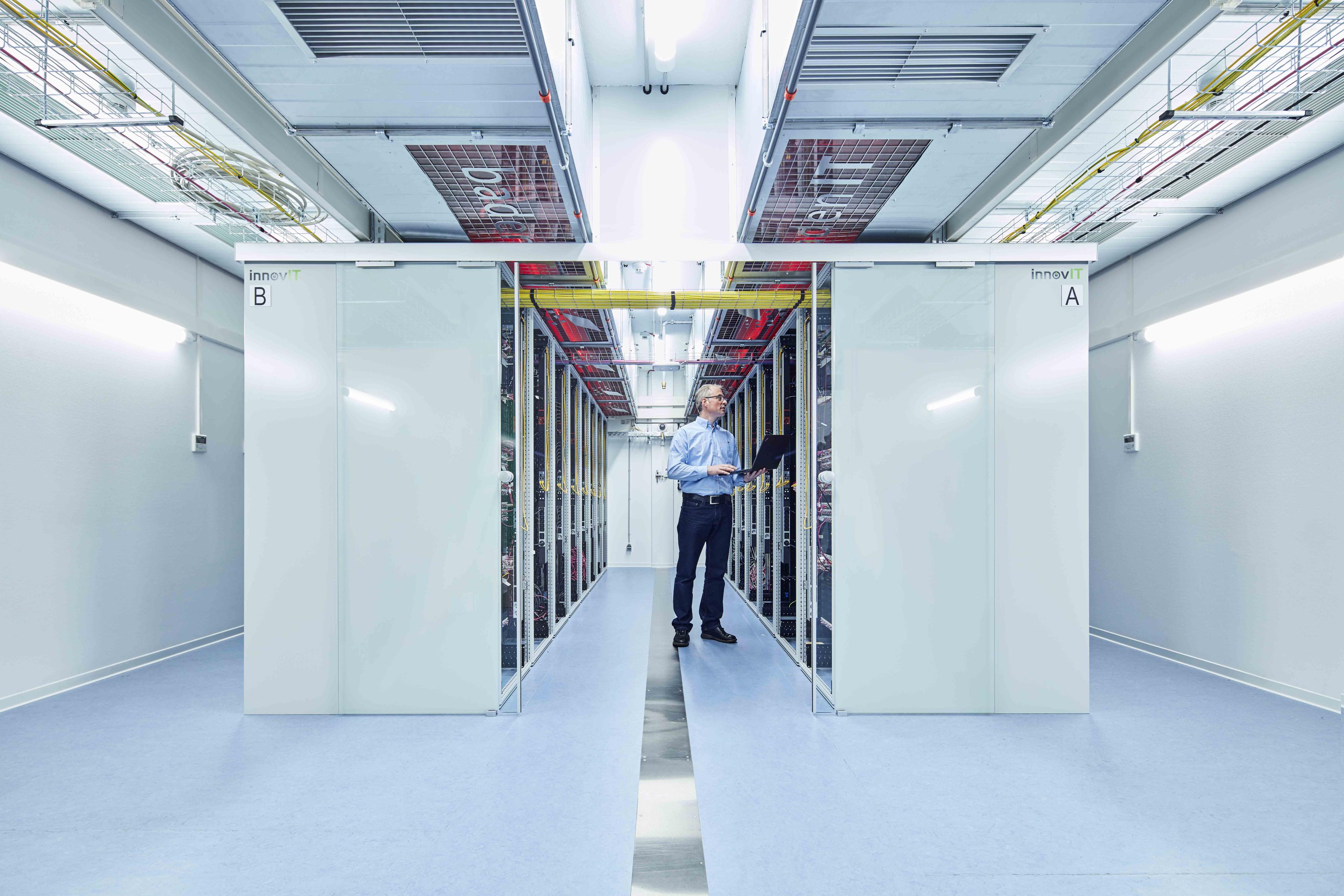 A person with a laptop stands in a corridor between two rows of server racks in a data center, surrounded by cables and electronic equipment.