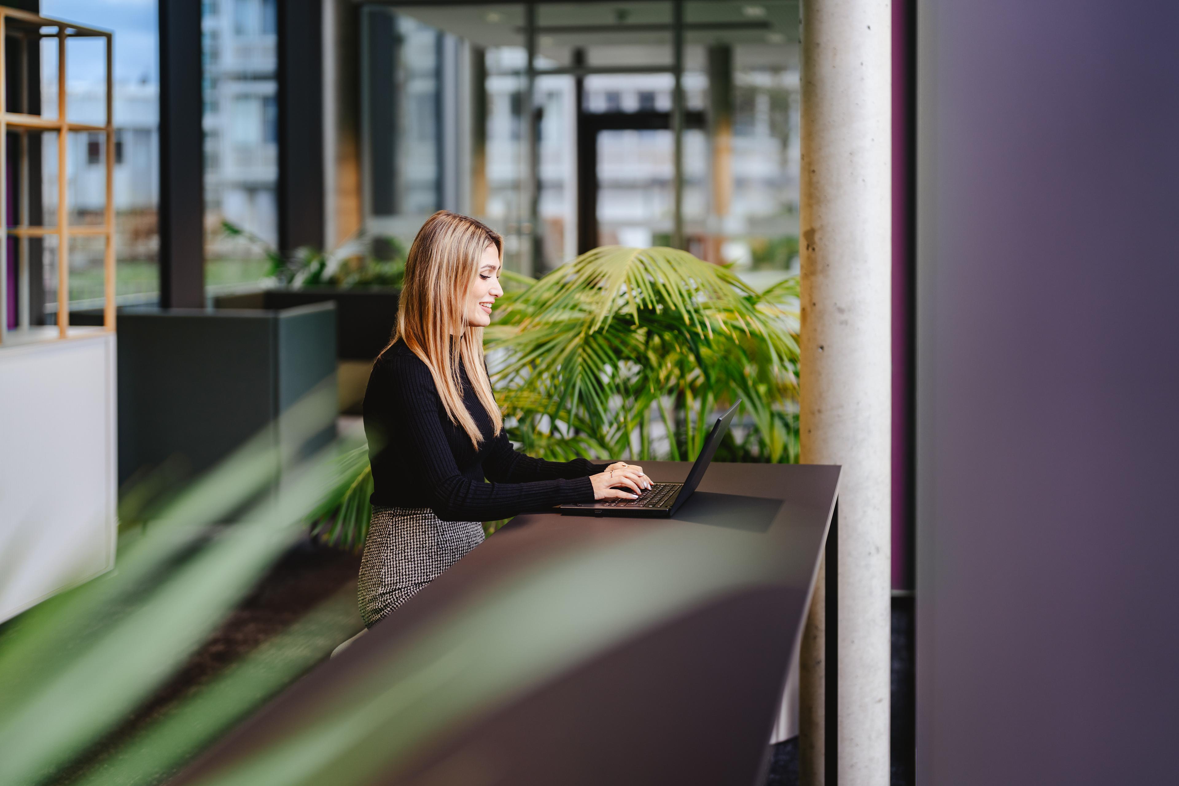 Eine Frau mit langen blonden Haaren sitzt an einem schwarzen langen Tisch am Laptop. Hinter ihr befinden sich viele Zimmerpflanzen, das Interieur ist modern und in schwarz weiß mit Holz gehalten.